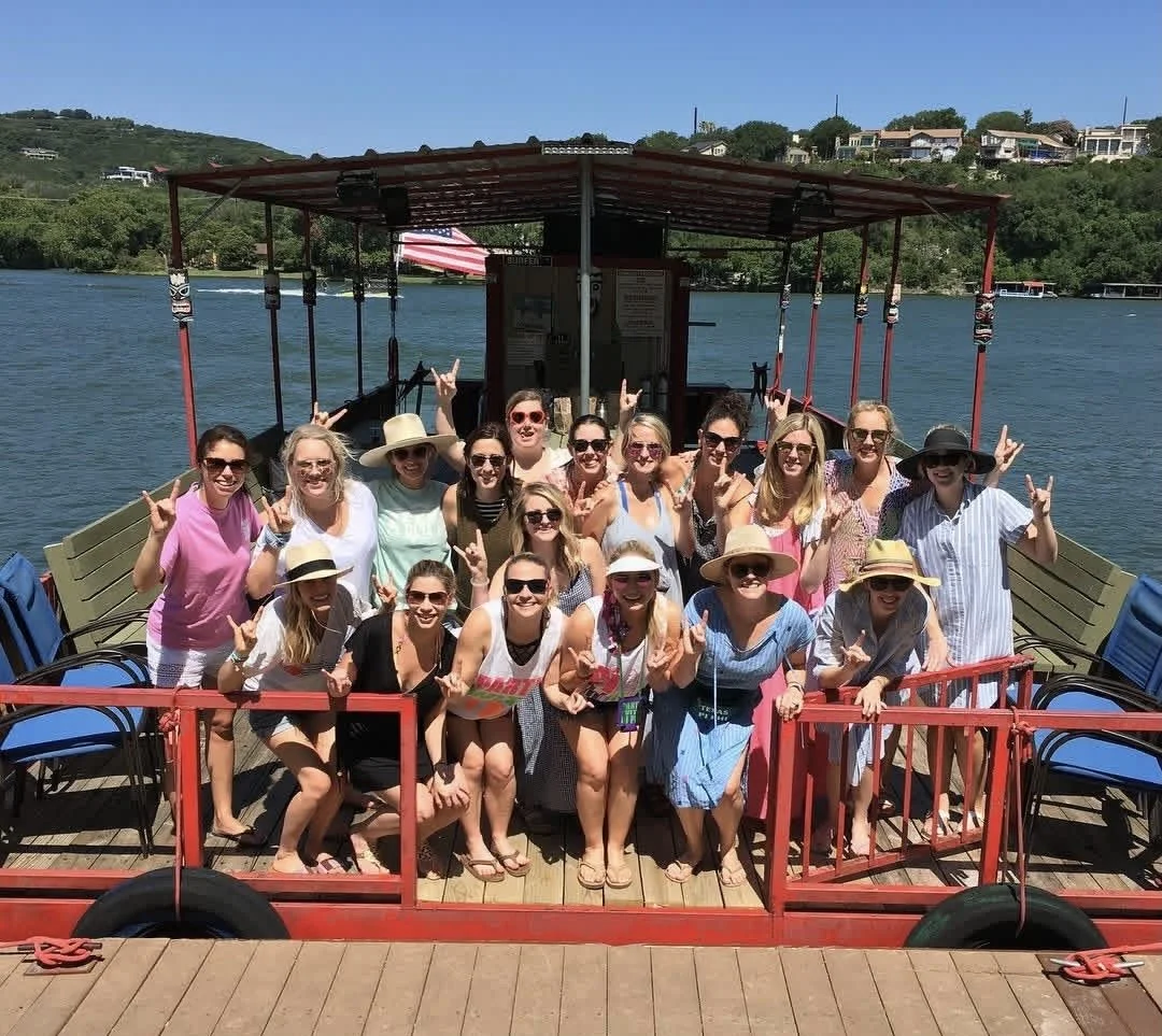 A group of women on a boat, posing for a photo with many making the 'rock on' hand gesture, on a sunny day with water and greenery in the background.