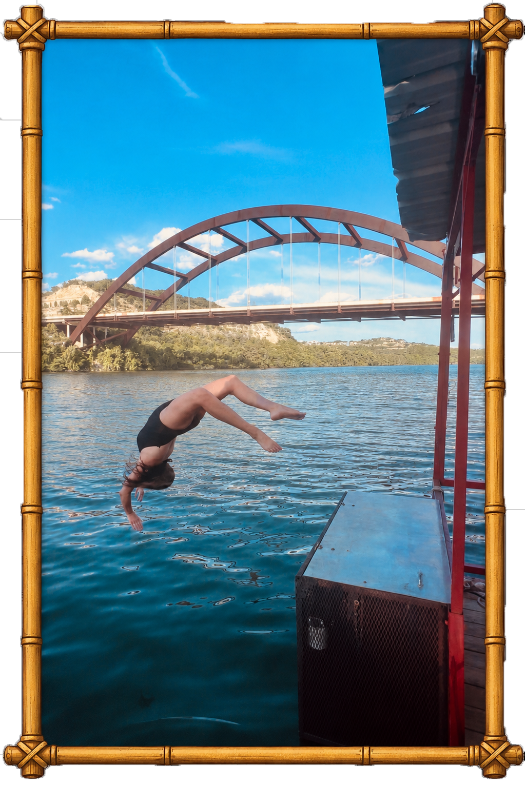 A person in a black swimsuit diving off a dock into a body of water with a bridge and hillside in the background under a blue sky with clouds.