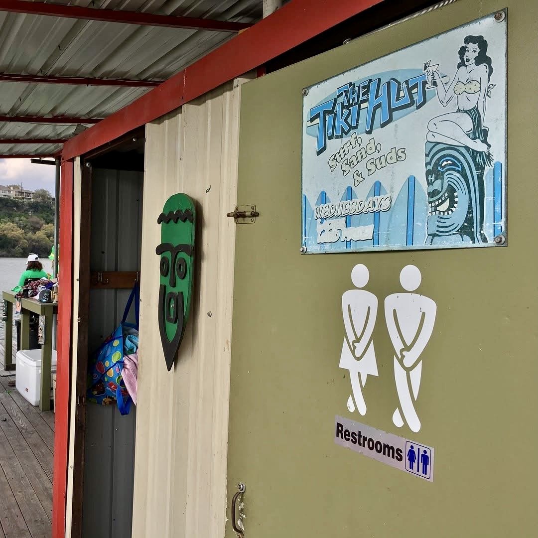 A sign indicating restrooms near a dock or outdoor area with people fishing. The sign features a retro-style illustration of a woman in a yellow top and striped skirt holding a drink, and a playful tiki-style face mask hanging on the wall.