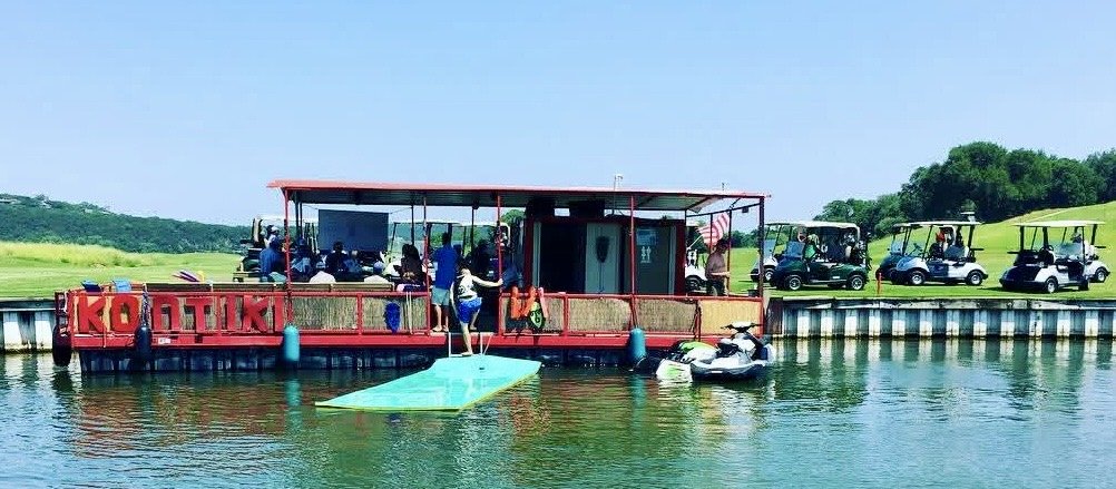 A floating golf cart bar on a lake with several people enjoying drinks, with golf carts parked on the grass nearby.