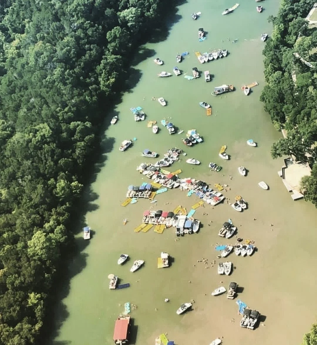 Aerial view of a boat dock on a river, with many boats moored along the shoreline and some boats dispersed in the water, surrounded by dense green trees.