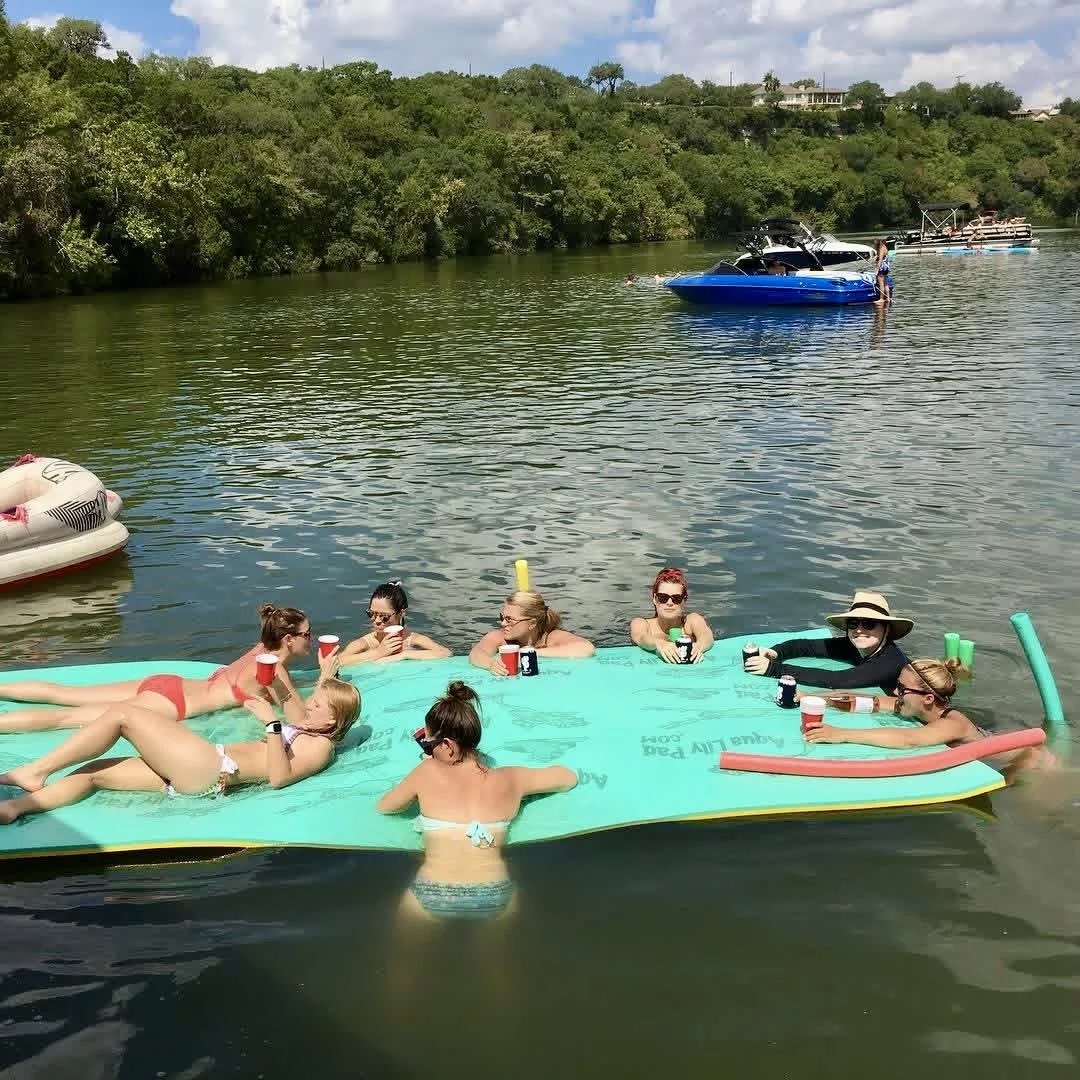 Group of women in swimsuits relaxing on an upside-down float with drinks, near a shoreline with trees, at a lake during daytime.