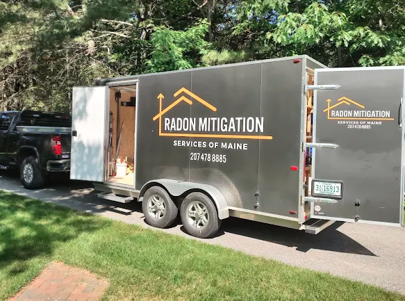 A black trailer truck with the words "Radon Mitigation" and "Services of Maine" written on the side, parked on the street with trees and a grassy area in the background.