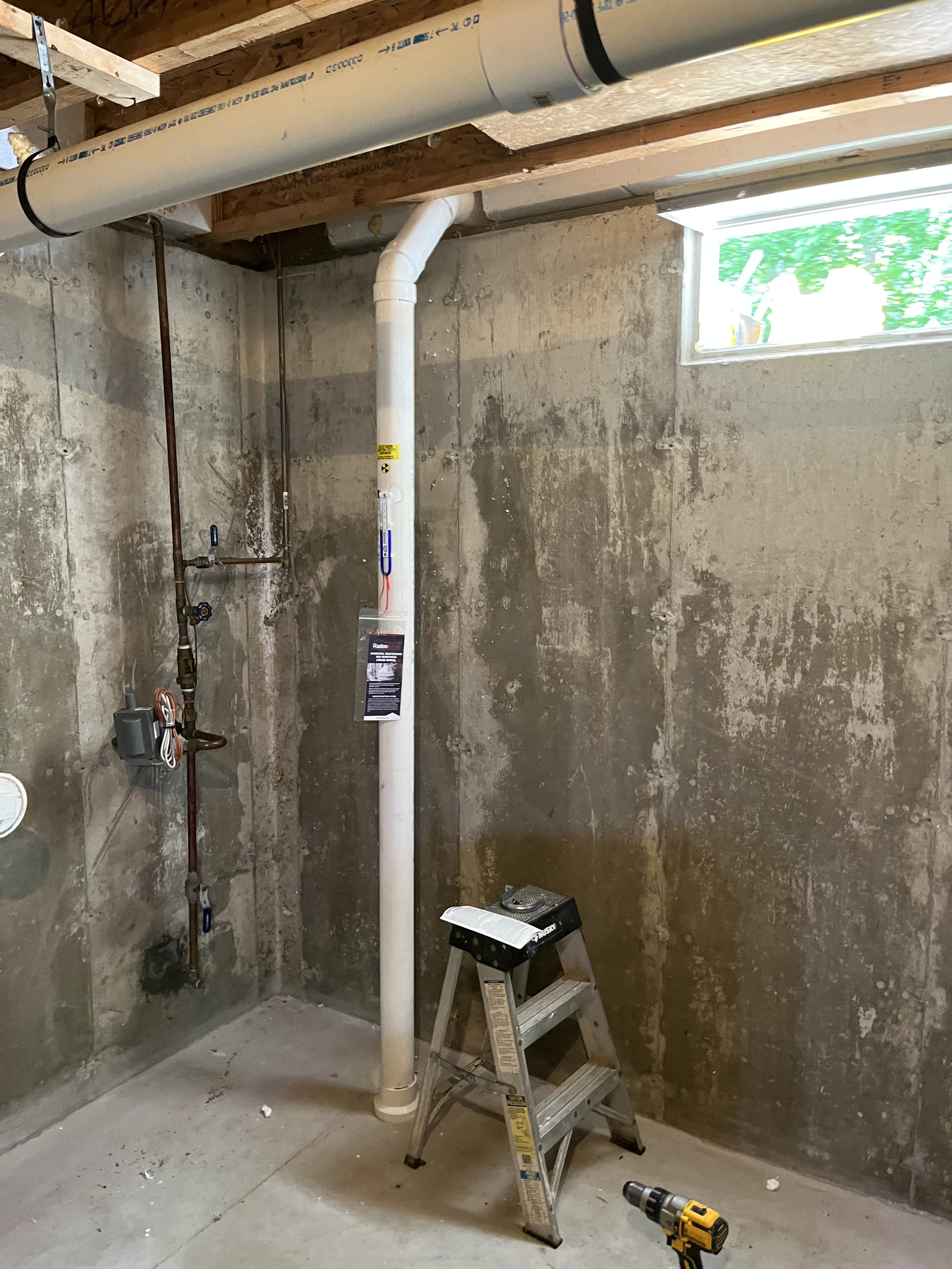Basement with exposed concrete walls, a small window, a white plastic pipe running vertically, a step ladder, a cordless drill, and construction tools.