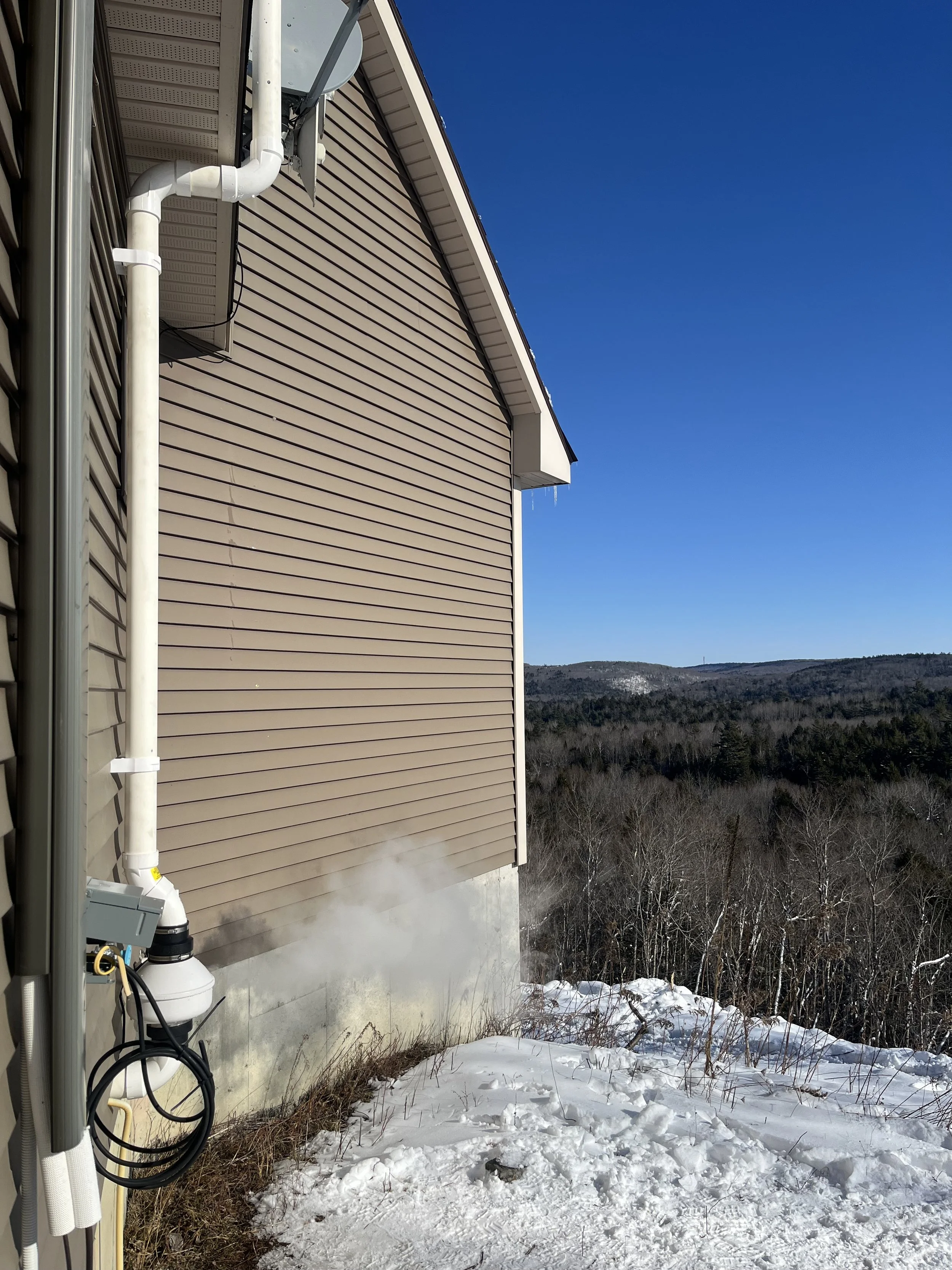 Side of a house with beige vinyl siding and white trim, with a vent pipe and electrical box near the foundation. Steam or smoke is rising from the ground near the house. Snow covers the ground, with a forested hillside in the background under a clear