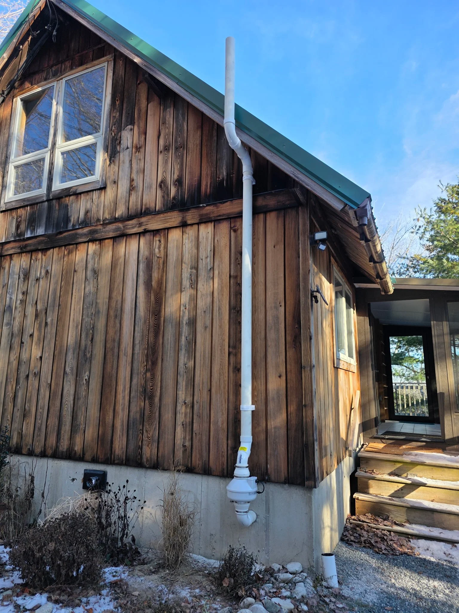 ABandoned house with wooden exterior, a drainpipe, and stairs leading to a back door, with plants and rocks at the base.