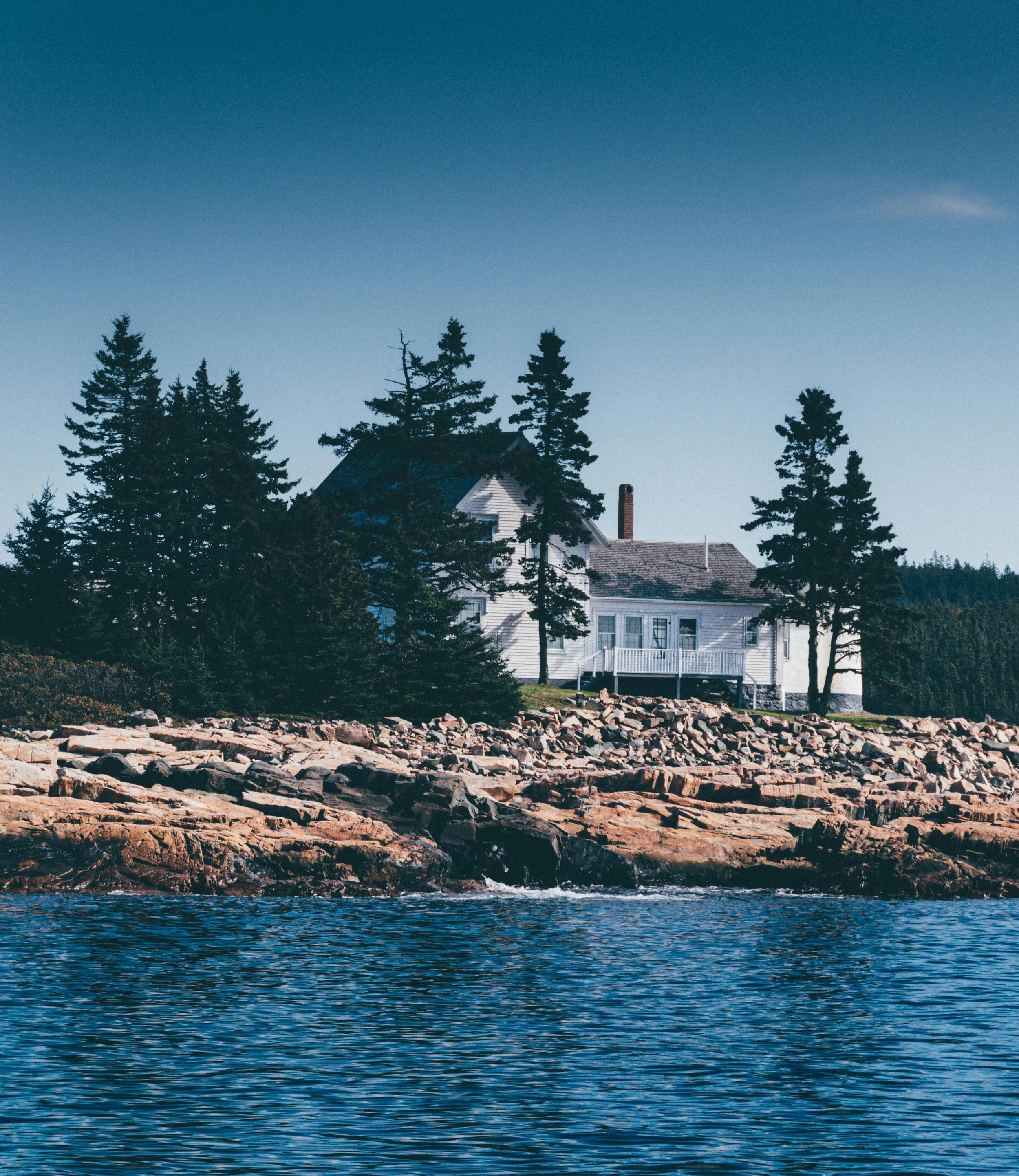 A white house with a deck stands on a rocky shoreline, surrounded by tall pine trees, with a body of water in the foreground and a clear blue sky above.