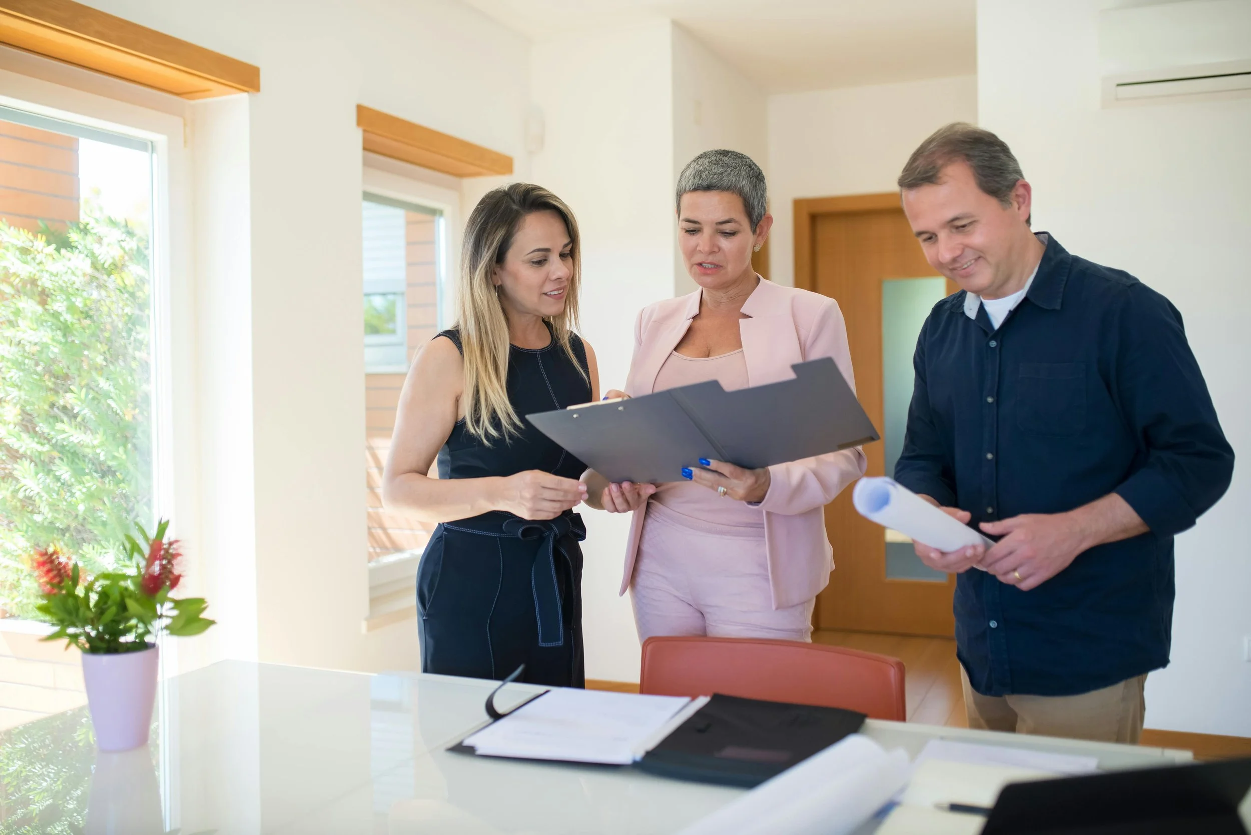 Three people standing around a table, reviewing documents in a bright room with large windows and potted plant.