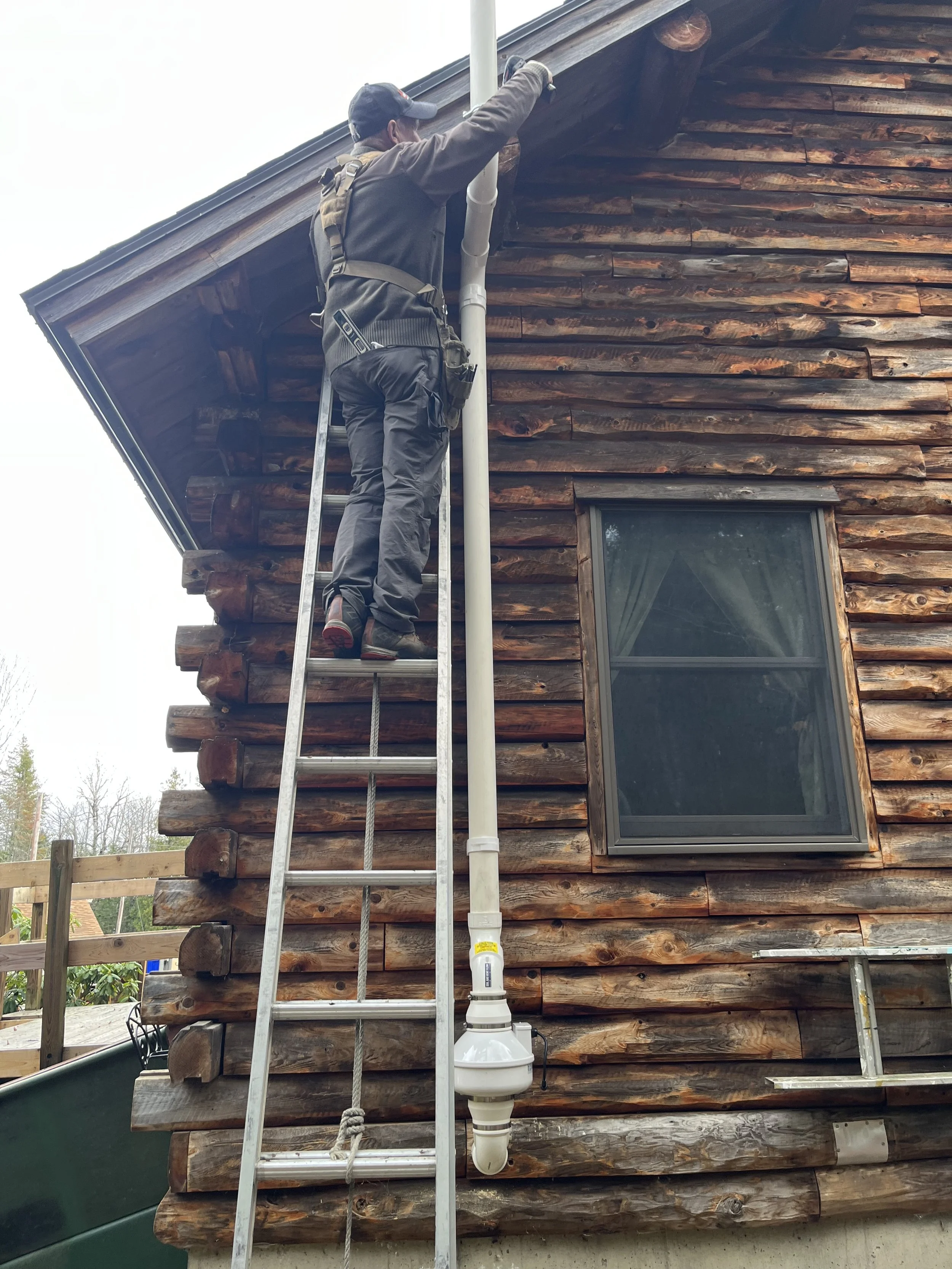 A man in dark clothing and a baseball cap working on the exterior of a wooden house using a ladder and safety harness.