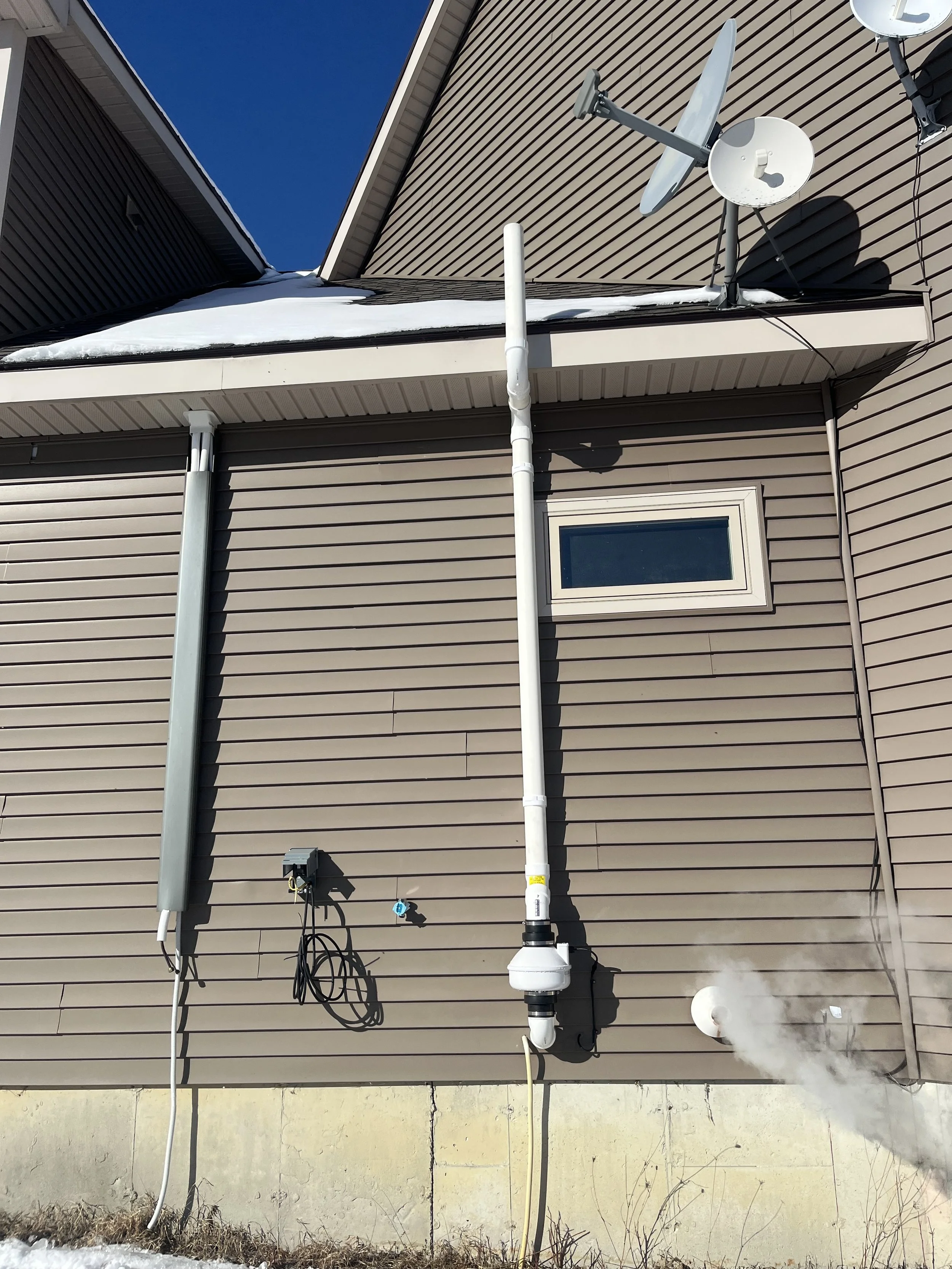 Back of house with satellite dishes, spray foam insulation, and utility connections, with snow on roof and blue sky.