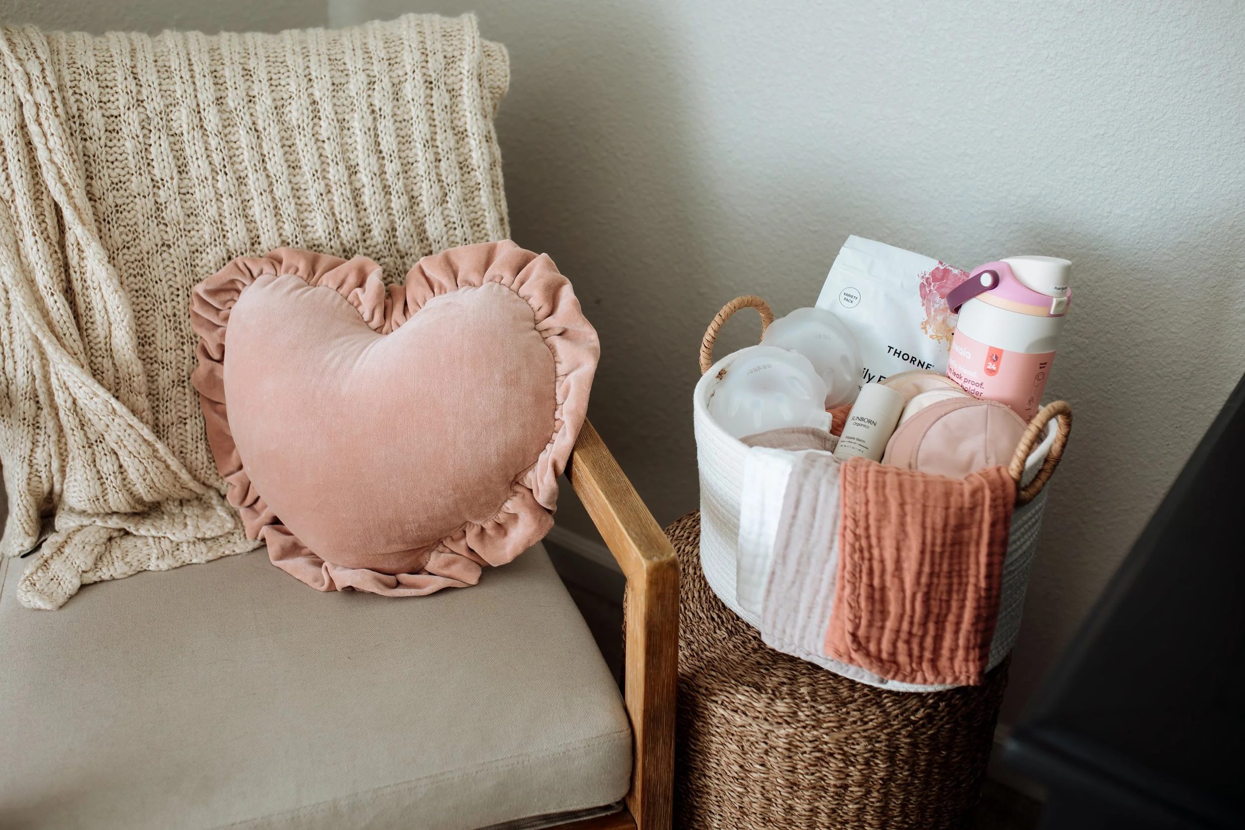 A beige armchair with a chunky knit blanket draped over the backrest, and a pink heart-shaped velvet cushion on the seat. To the right, a basket holds postpartum products, a pink water bottle, and folded towels.