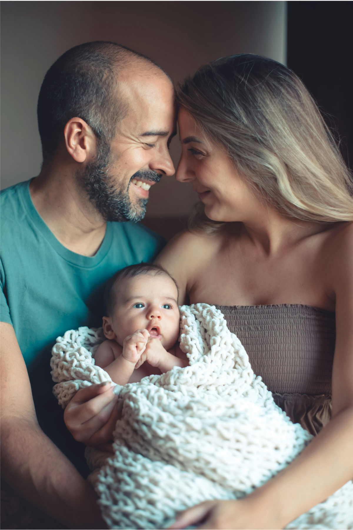 A happy family with a man, woman, and a newborn baby. The man and woman are smiling and touching foreheads, holding the baby wrapped in a cozy blanket.