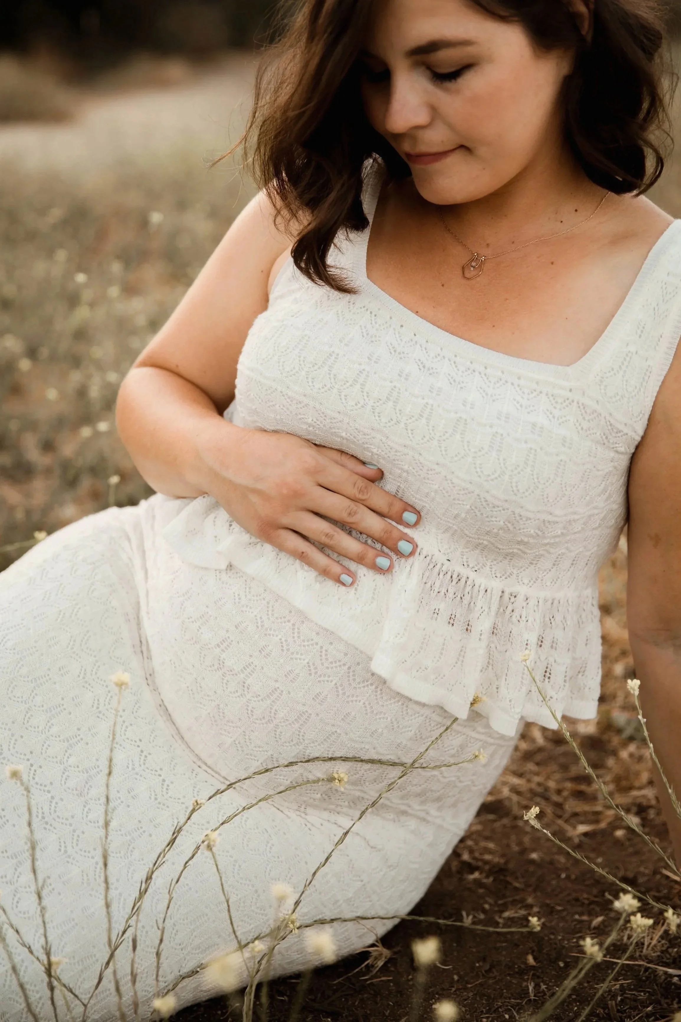 A woman wearing a white lace dress and necklace, squatting on the ground outdoors, surrounded by dried plants and soil.