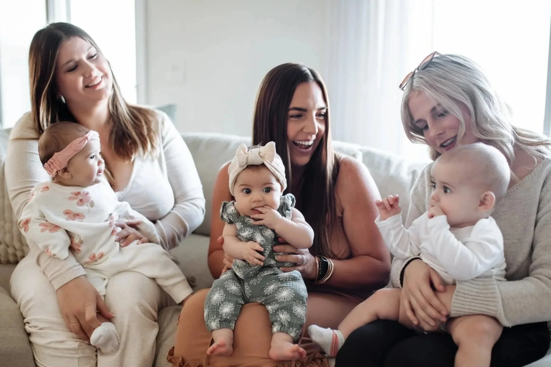 Group of three women with three babies sitting on a beige couch in a bright room with white curtains. The women are smiling and engaging with the babies.