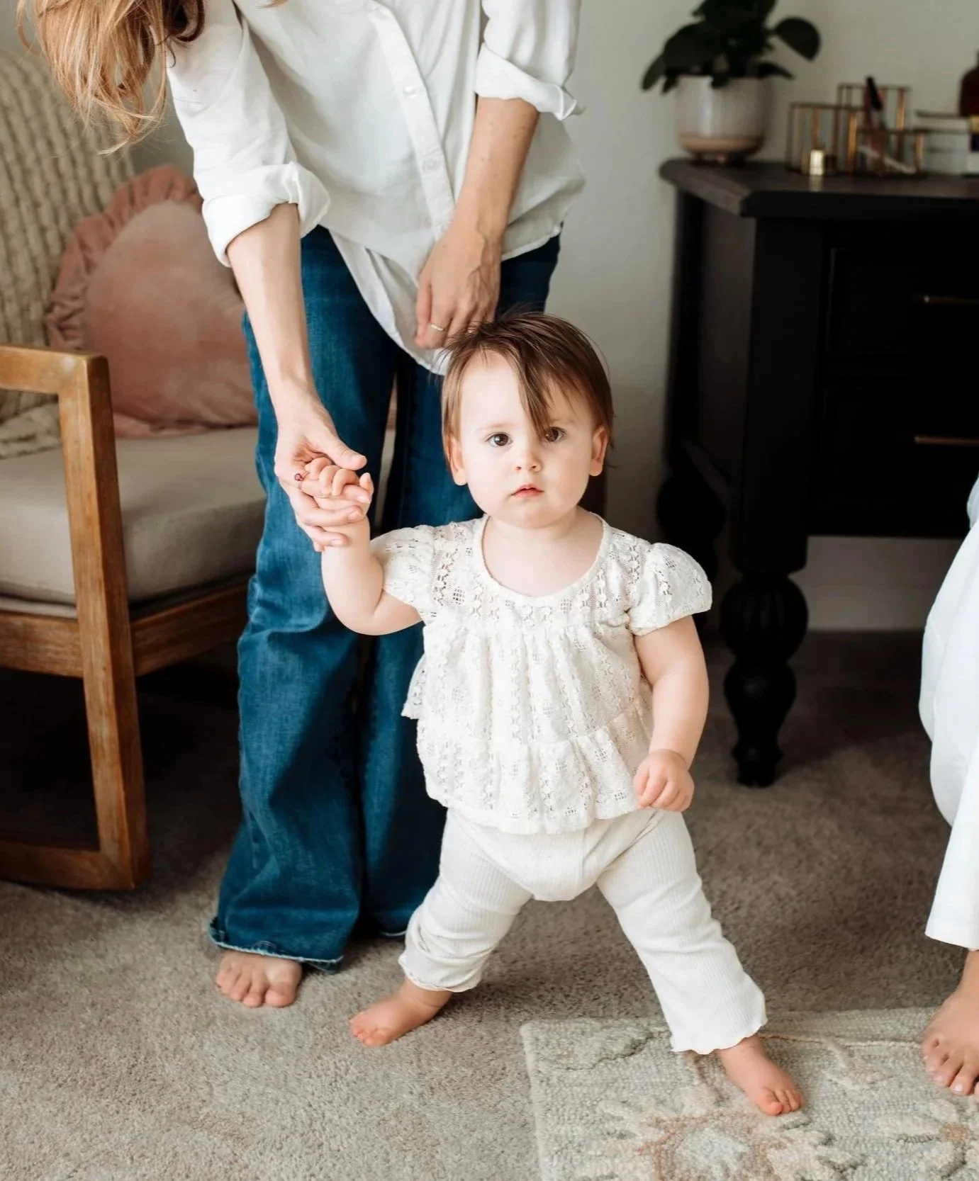 A young girl with brown hair and a white lace top standing on a beige carpet, holding hands with an older person. The girl has a serious expression and is in a slight squat.