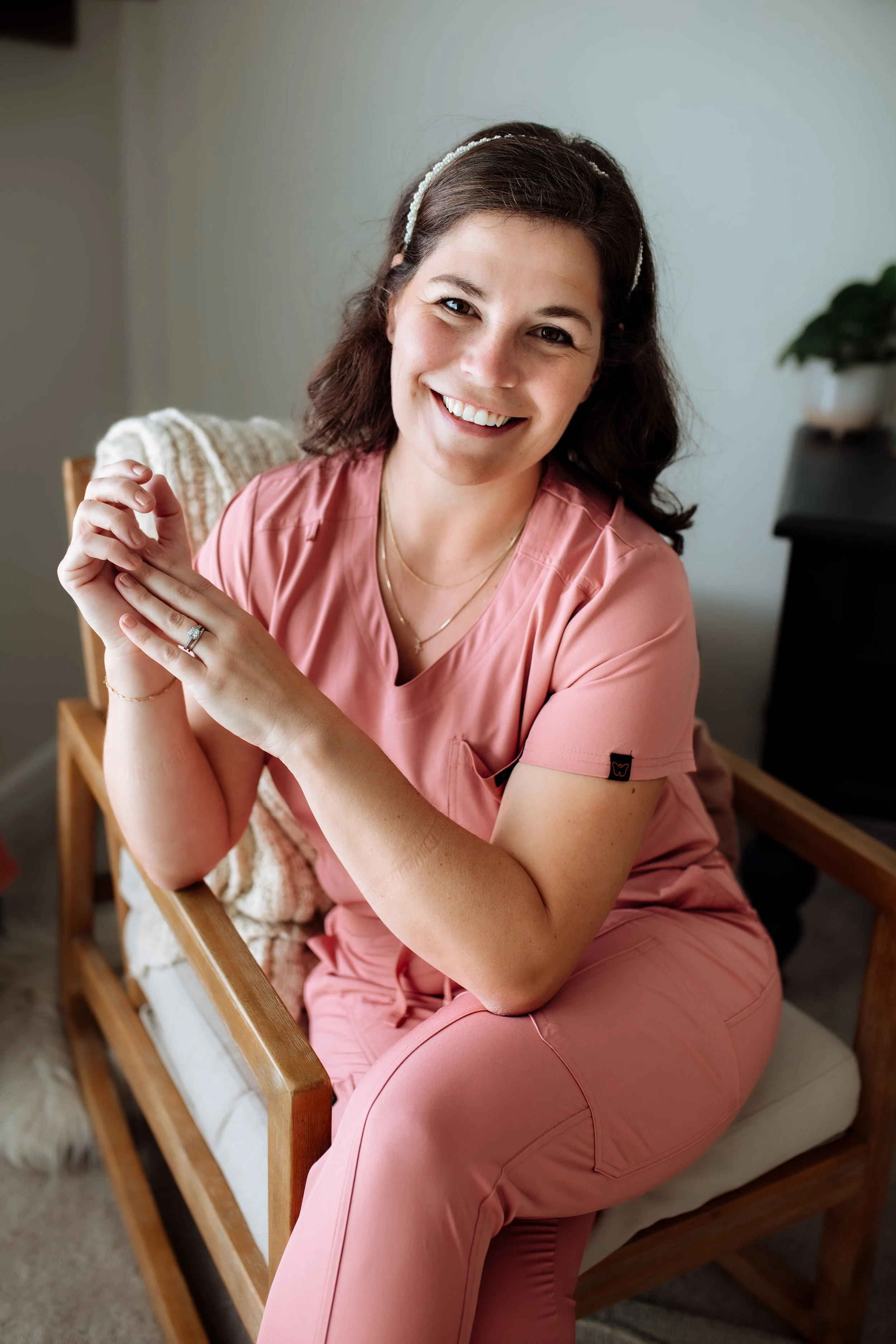 A woman with brown hair and a headband, smiling and sitting on a wooden bed frame with a blanket, wearing pink scrubs, in a well-lit room with a light-colored wall and a plant in the background.