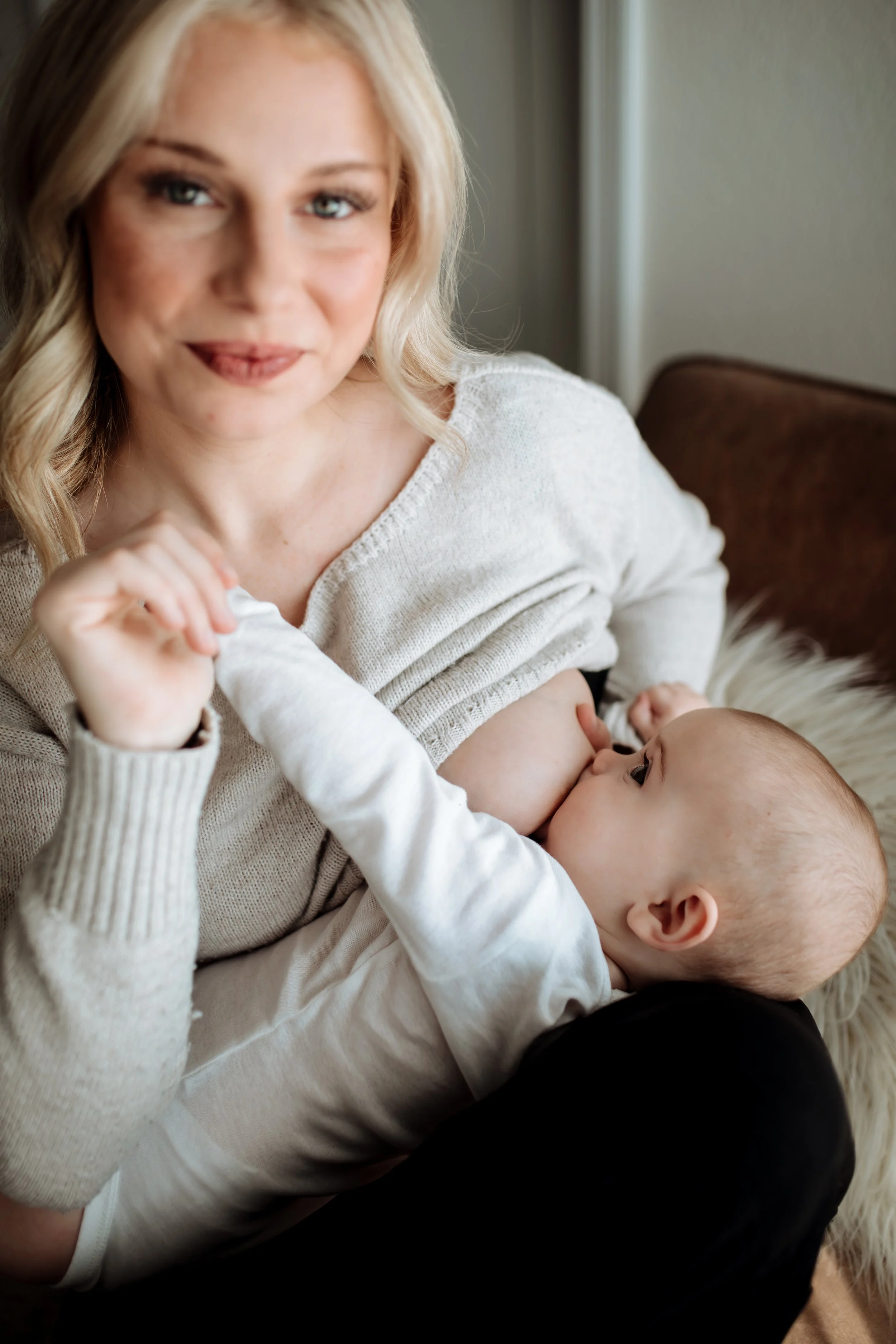 A woman with blonde hair, wearing a white sweater, breastfeeding a baby who is lying on her lap.