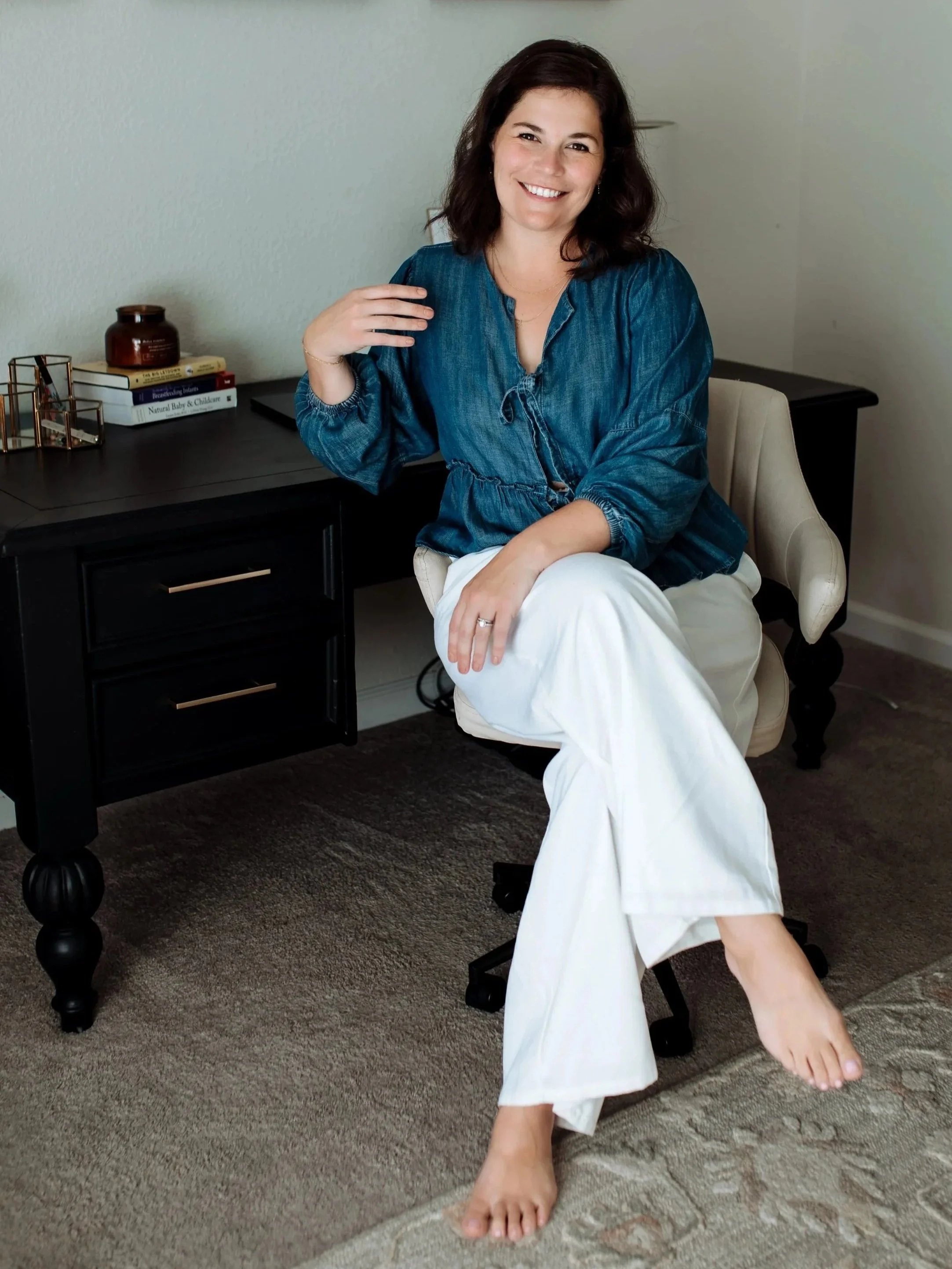 A woman with dark hair, wearing a teal blouse and white pants, sitting in a chair with one foot on the carpeted floor and the other extended, smiling at the camera in a room with a black desk, books, and decorative items.