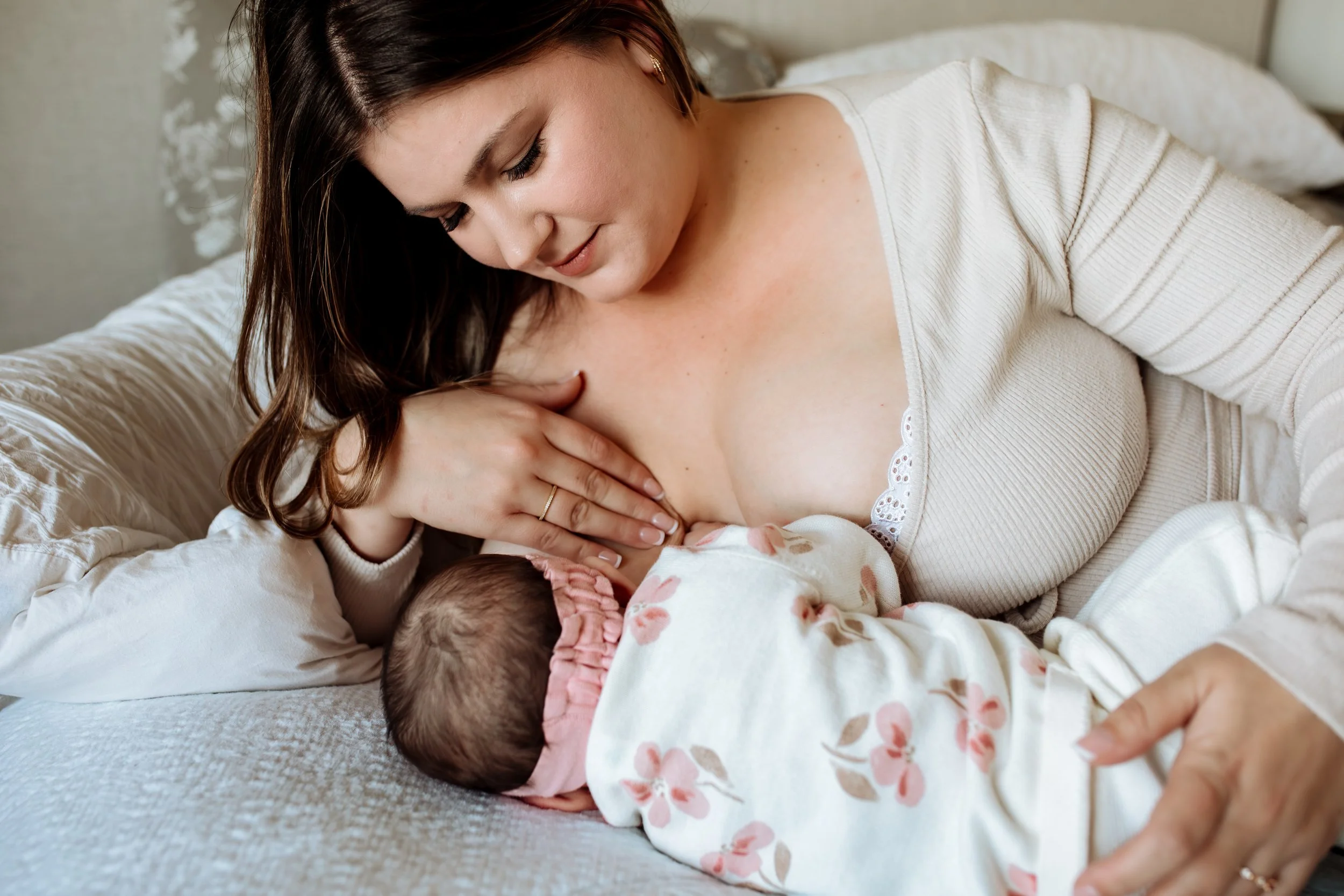 A woman with brown hair and light makeup nursing a newborn baby in bed.