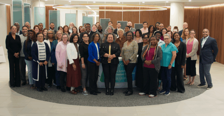 Group of diverse professionals gathered indoors for a photo, standing around a table with a banner, in a modern office lobby
