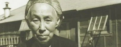 A black-and-white photo of an elderly Asian woman with a serious expression, standing outdoors in front of a house with a sloped roof and a rocking chair.