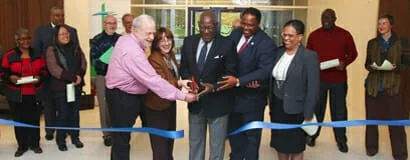 Group of diverse people at a ribbon-cutting ceremony, with a man in a pink shirt handing an award or certificate to a man in a suit.