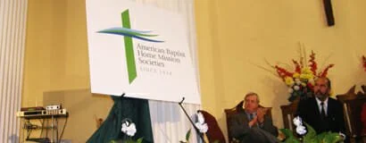 A sign for the American Taping, Finishing, and Onsite Association with the organization's logo, placed on an easel. In the background, two men are seated at a table with a floral arrangement.