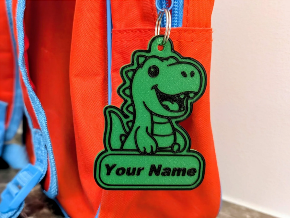 A green dinosaur-shaped tag with a cartoon face and the words 'Your Name' attached to a red and blue backpack.