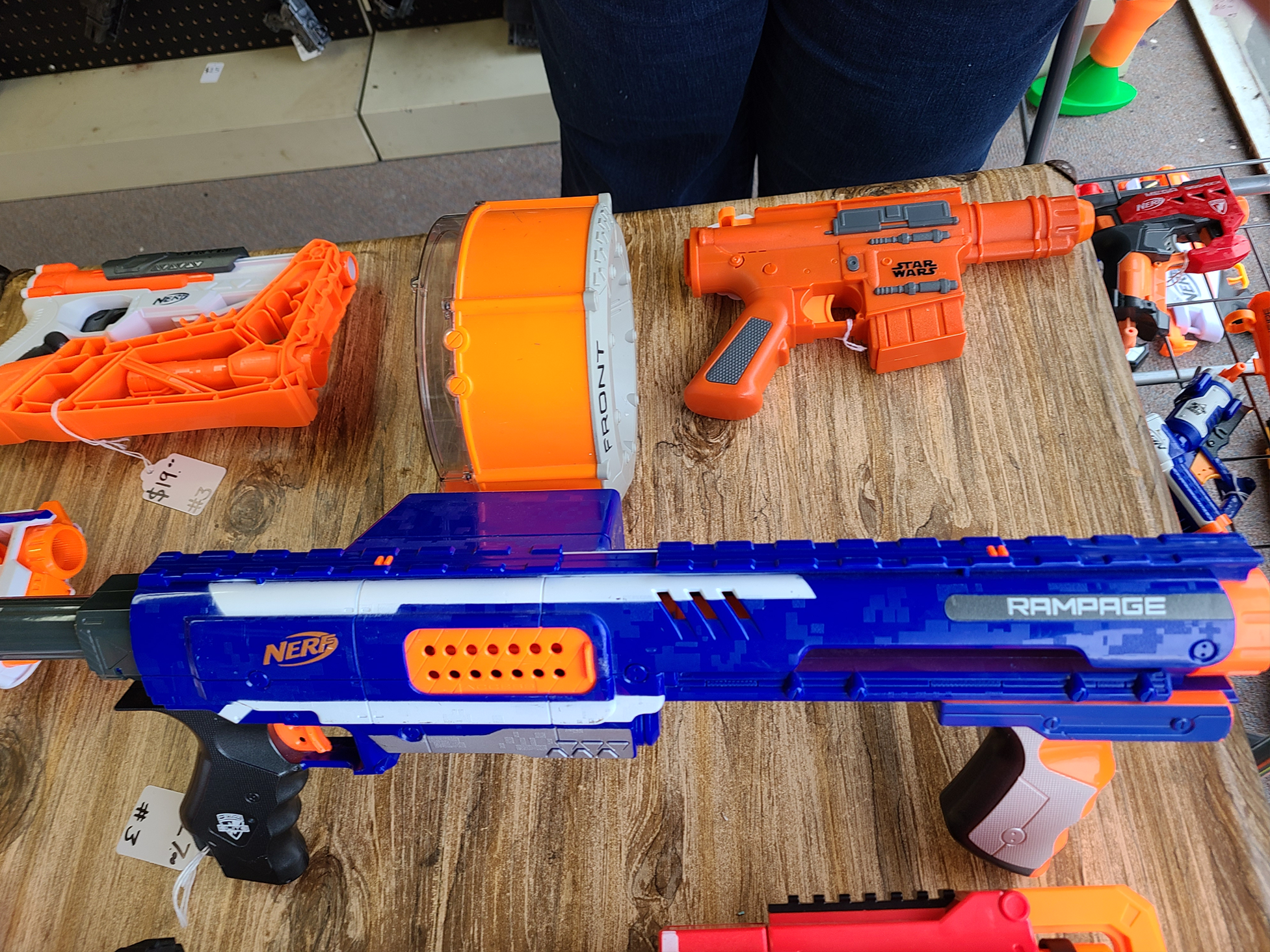 Display of toy Nerf guns on a wooden table, including a large blue Nerf Rampage, an orange Star Wars-themed gun, and other colorful Nerf blasters.