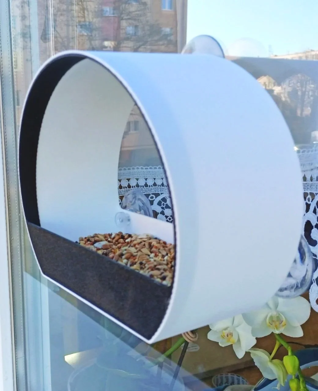 A modern 3D printed white pet feeder, filled with bird seed, suction-cupped on the outside of a window with a lace curtain and orchid flowers nearby.