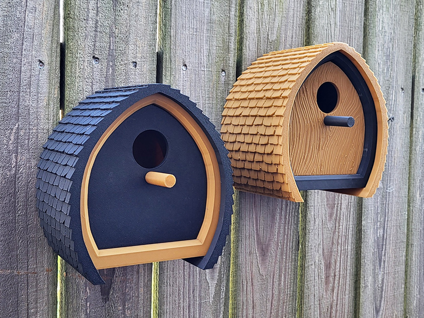 Pair of modern 3D printed birdhouses mounted on a wooden fence, one black and the other natural wood-colored.