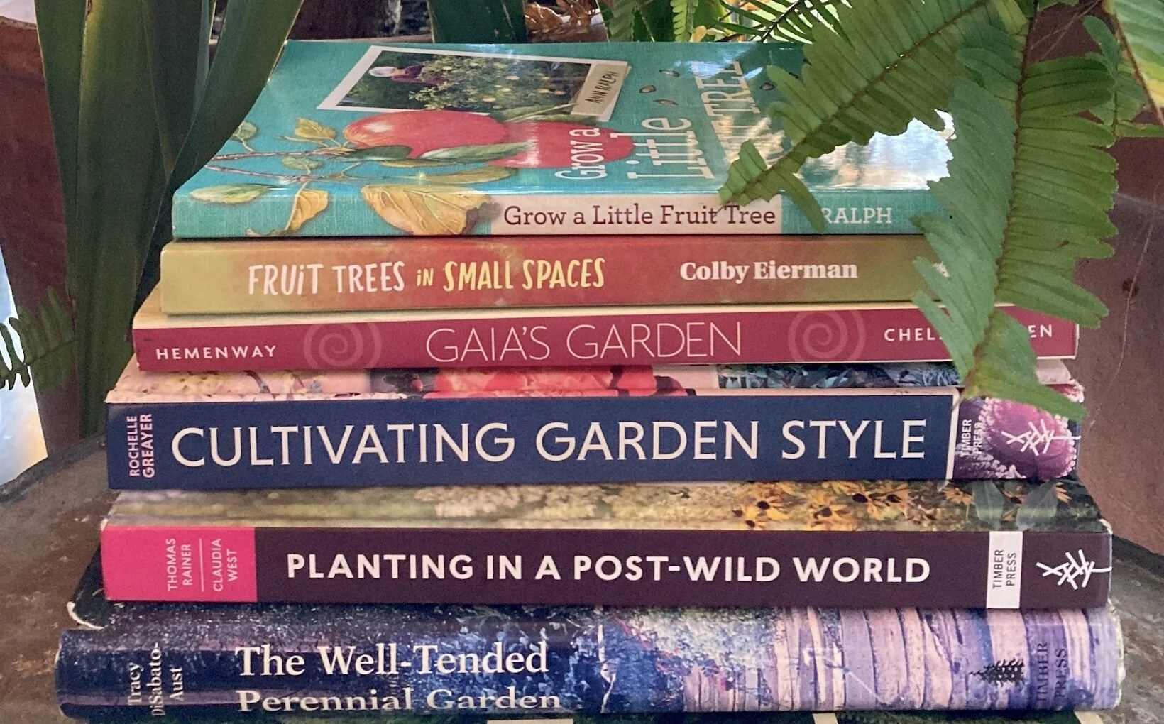 Stack of gardening books on a wooden surface, surrounded by green foliage.