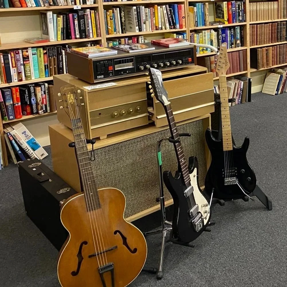 Three guitars, a black amplifier, and a vintage radio on a wooden table in front of a bookshelf filled with books in a room.