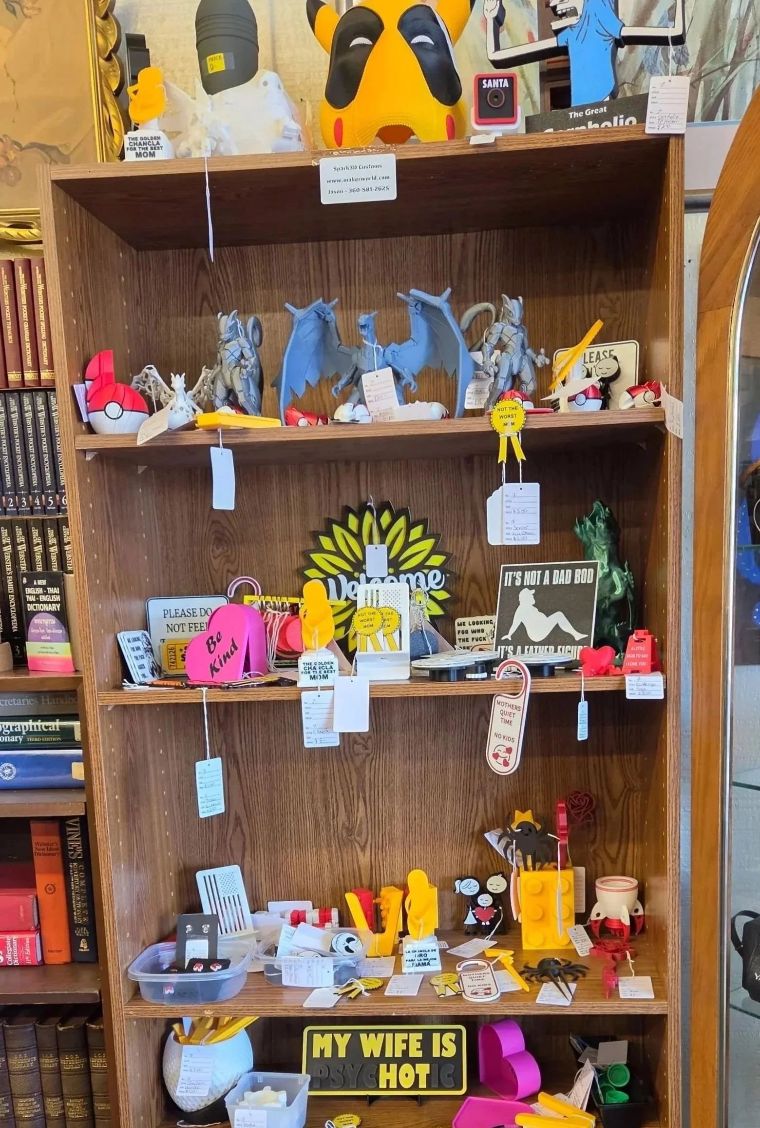 A wooden shelf filled with various 3D printed novelty and humorous items such as signs, figurines, and magnets, including a sign that reads 'MY WIFE IS PSYCHOTIC' and other playful objects.