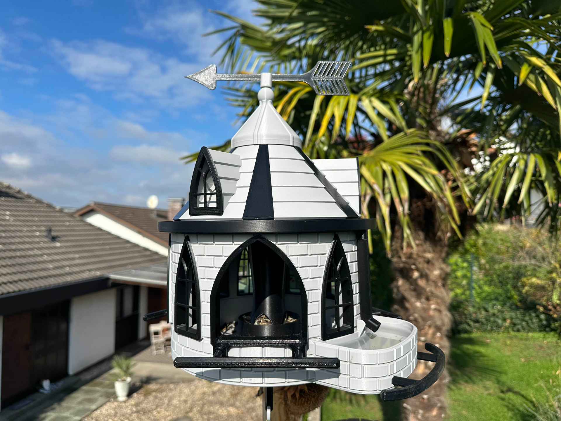 A decorative 3D printed birdfeeder shaped like a house with a silver weather vane on top and a water trough on the side, mounted on a pole outdoors during a sunny the day.