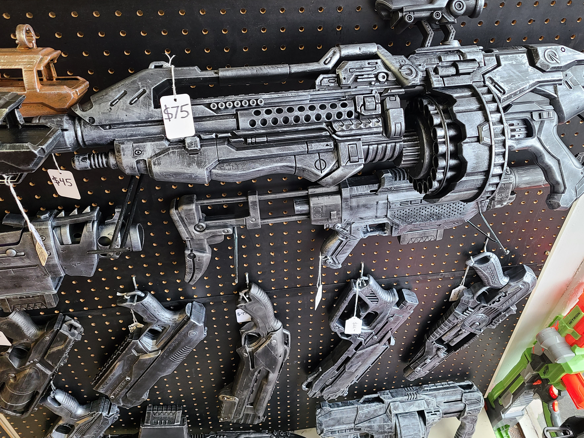 A large, futuristic-looking custom painted toy Nerf gun or sci-fi weapon with a black and gray color scheme hangs on a pegboard display at a store. The weapon has a detailed design with a drum magazine and a price tag showing $75.