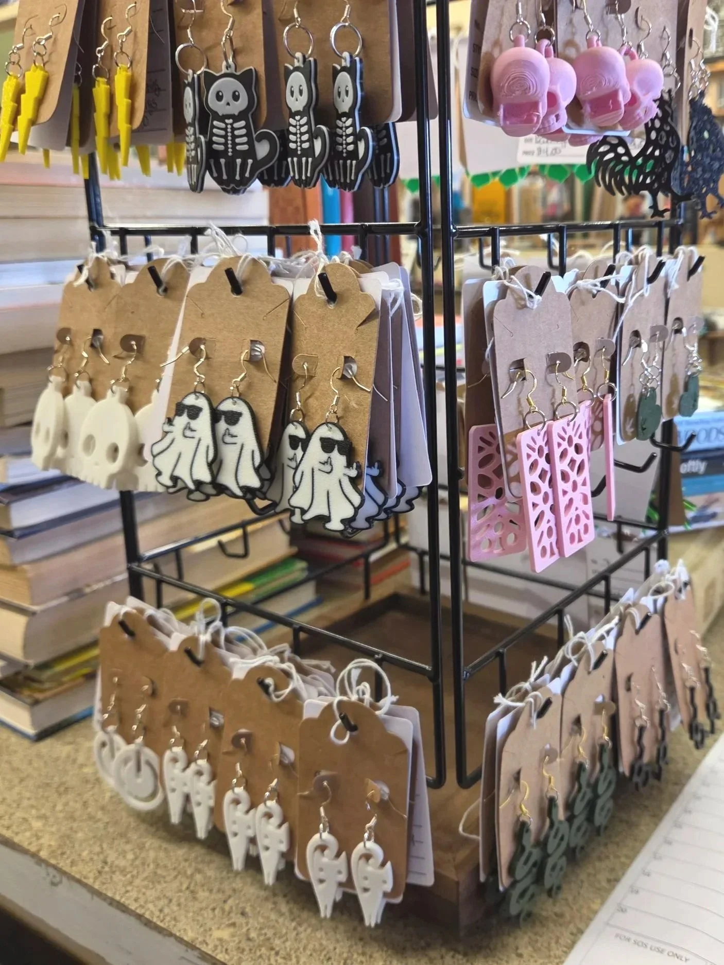 Assorted 3D printed earrings on display, including skeleton, ghost, pink skull, and abstract designs, on a multi-tiered jewelry stand in a store.