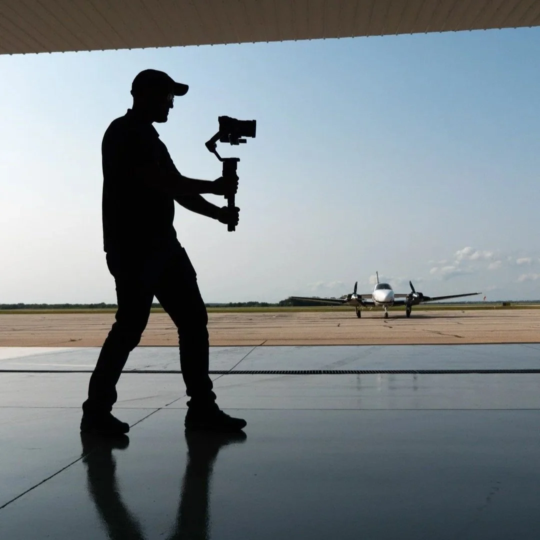 Silhouette of a person standing in front of an open hangar door, holding a camera stabilizer, with a plane on the tarmac outside.