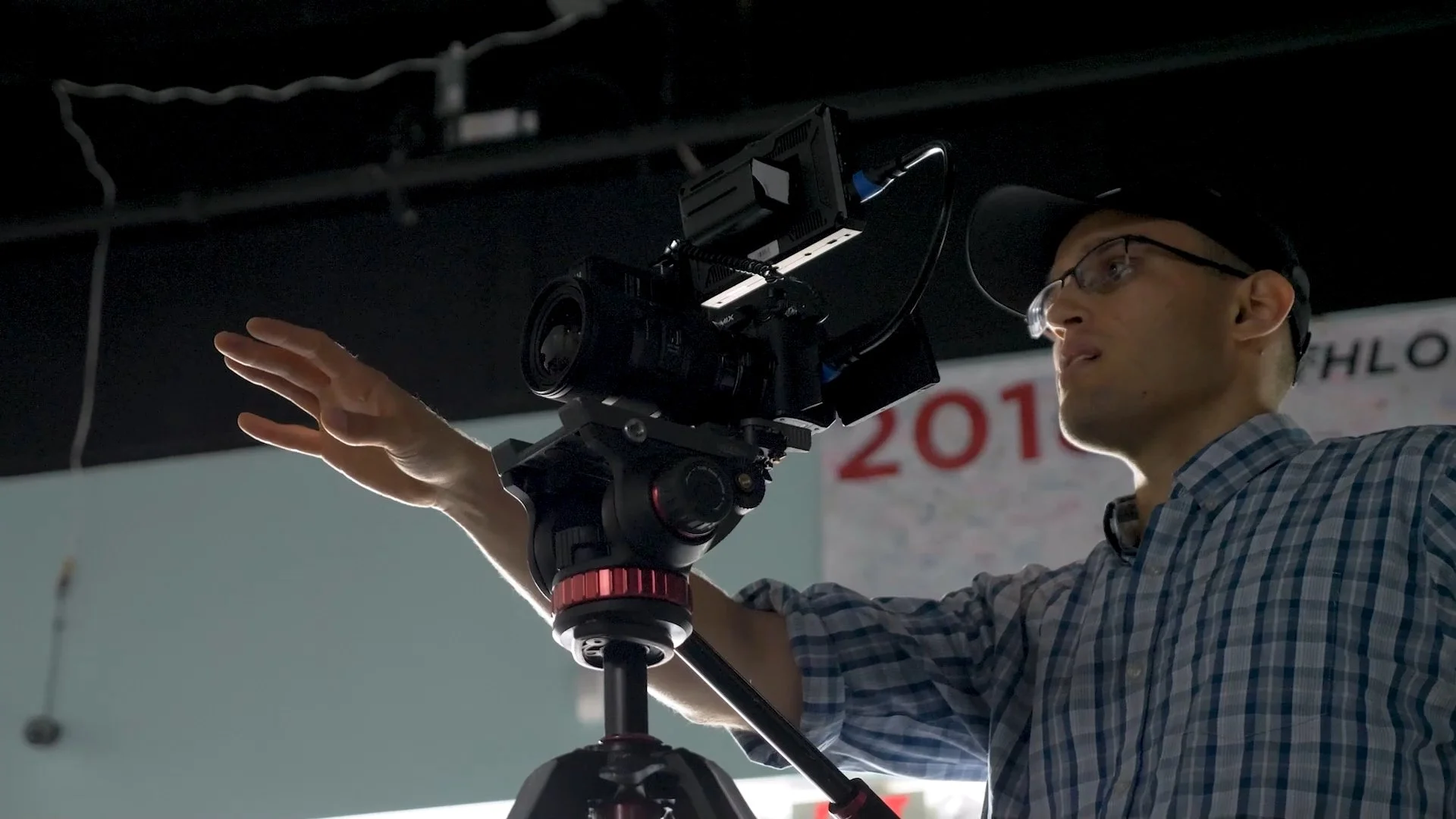 A man with glasses and a checkered shirt adjusting a professional camera mounted on a tripod, indoors with a partial sign in the background reading "201".