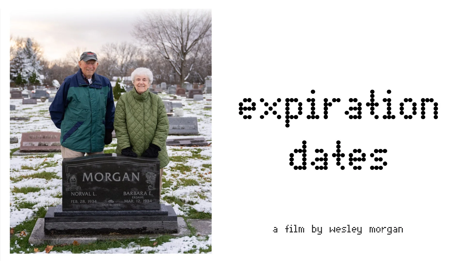 Two elderly people standing in a snow-covered graveyard, smiling at the camera. The man wears a cap, a blue, green, and black jacket, and beige pants, while the woman wears a green quilted coat and black gloves. A gravestone in front of them reads 'Morgan' with names and dates.
