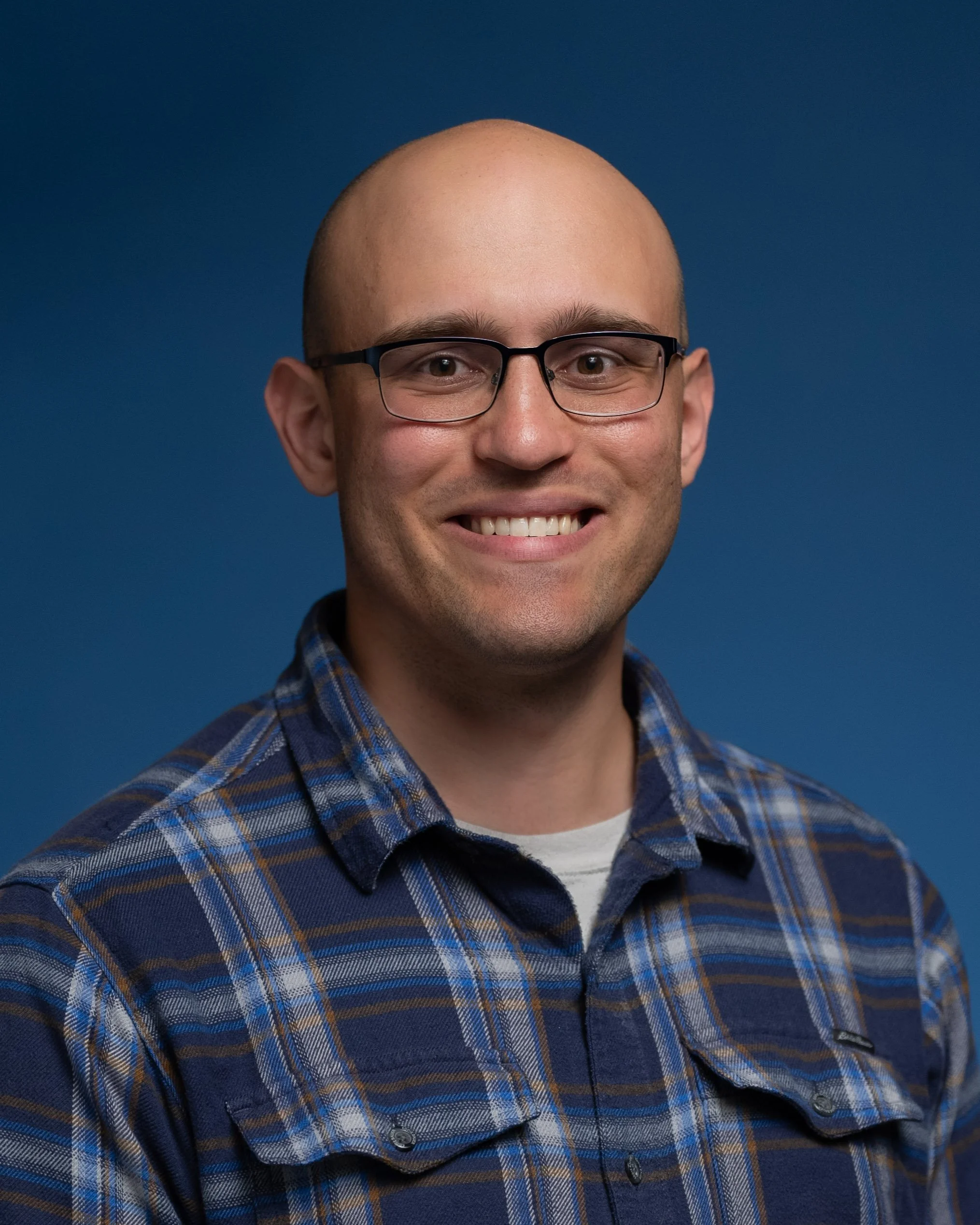 A man with glasses and a smile, wearing a blue plaid shirt, standing in front of a blue background.