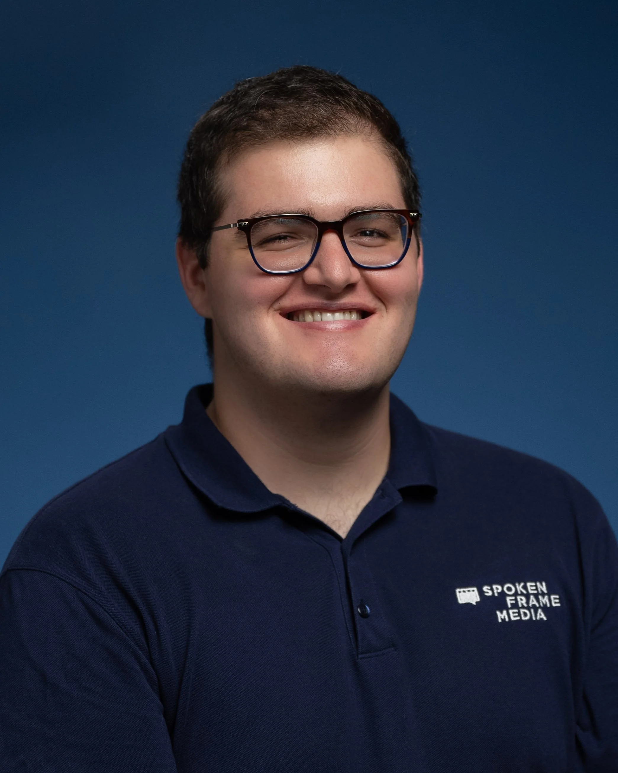 Young man with short brown hair and glasses smiling, wearing a navy blue polo shirt with a logo on the chest that says 'Spoken Frame Media' against a blue background.