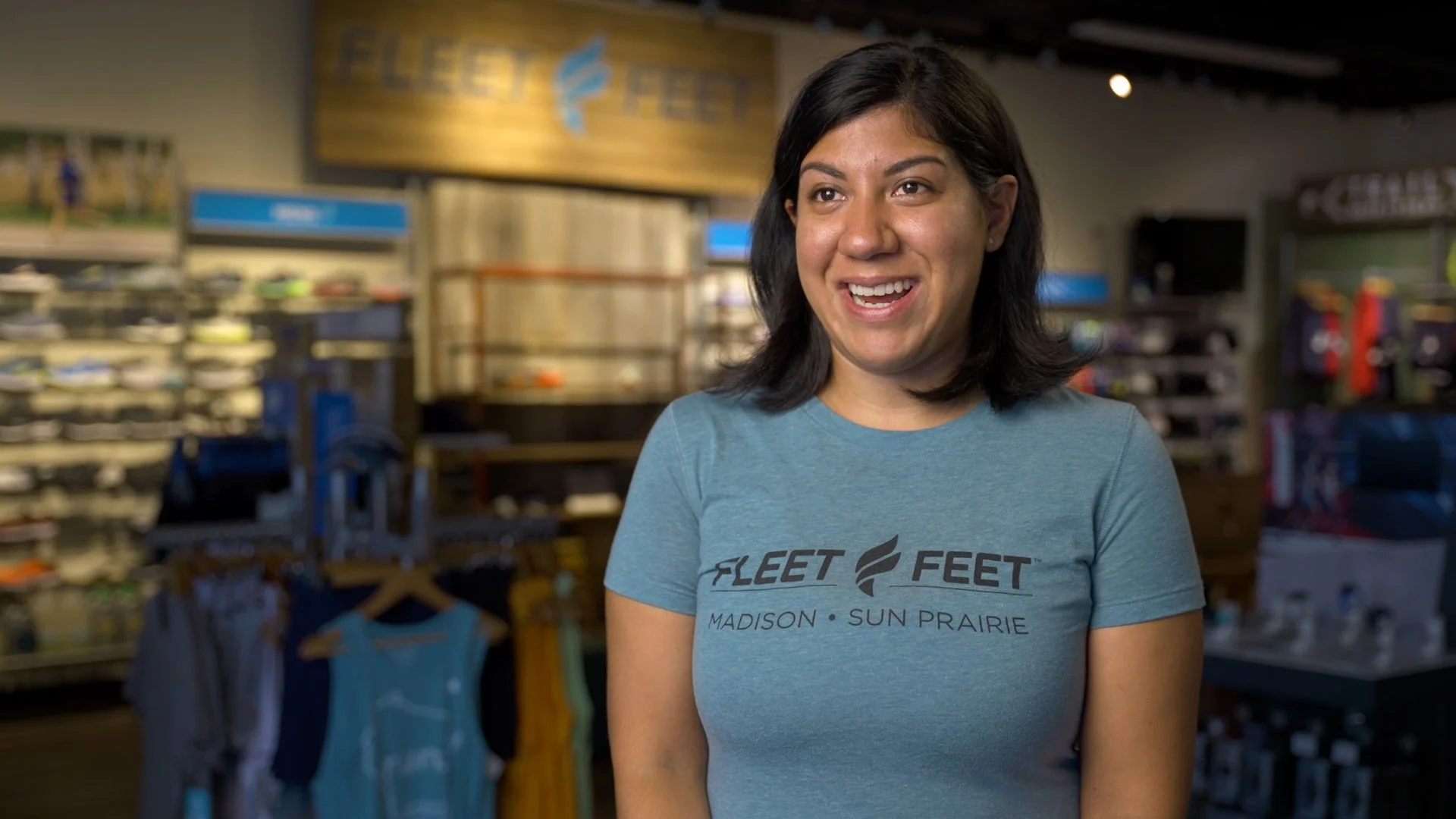 A young woman with dark hair wearing a blue T-shirt with the words 'FLEET FEET' and locations 'MADISON', 'SUN PRAIRIE' printed on it, smiling in a sports store.