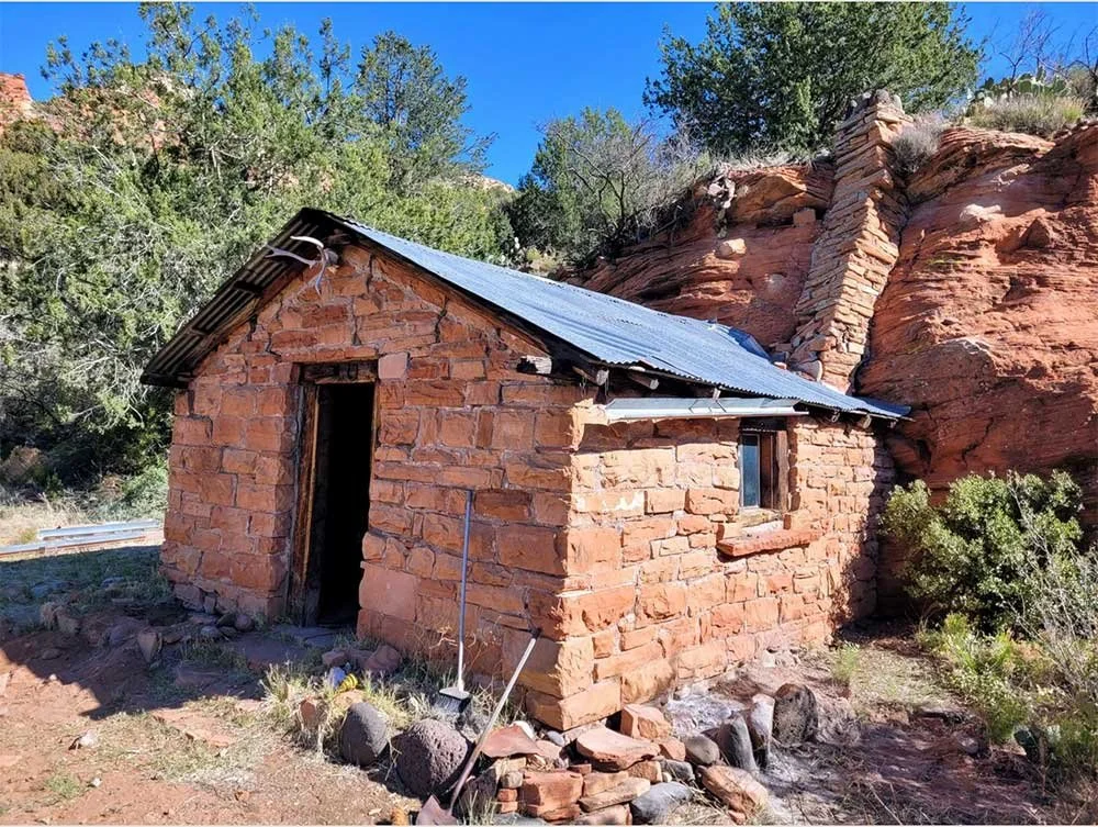 A small, historical brick cabin with a metal roof and a brick chimney, set against a red rock hillside with greenery.