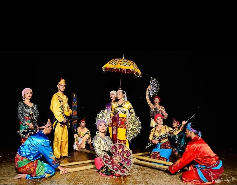 Traditional Indonesian performing arts with performers in colorful costumes and accessories, including umbrellas and fans, on stage.