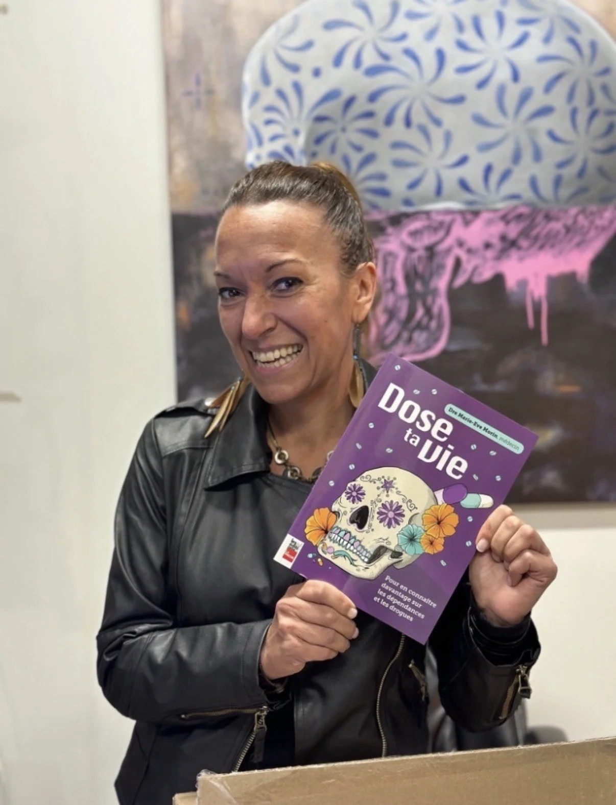 Marie-Eve Morin  tenant un livre intitulé 'Dose ta Vie' avec une illustration de crâne décoré de fleurs et de capsules de médicaments.