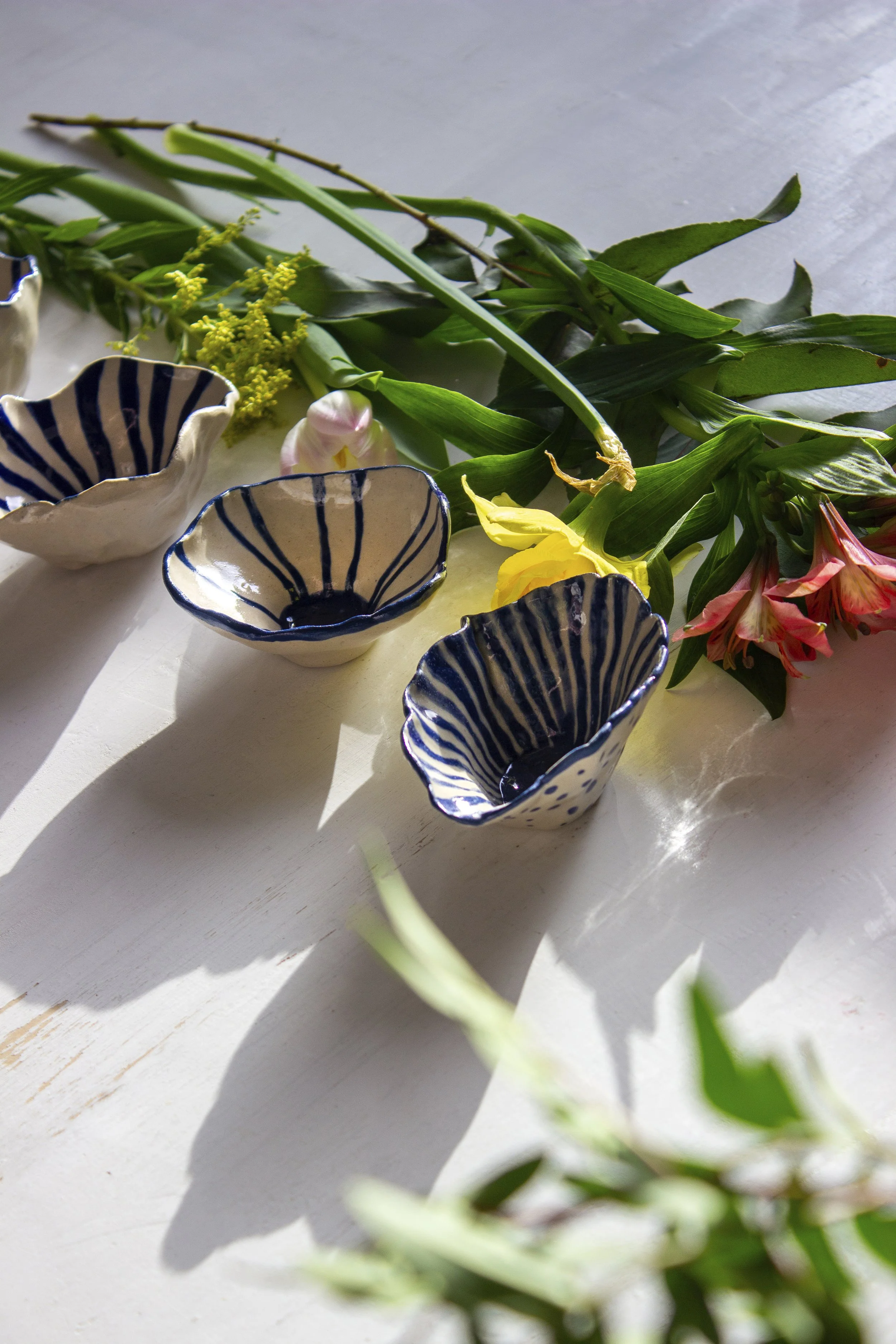 Siniraidallisia kuppeja pöydällä, niiden takana kukkia valkoisella tasolla auringonvalossa.