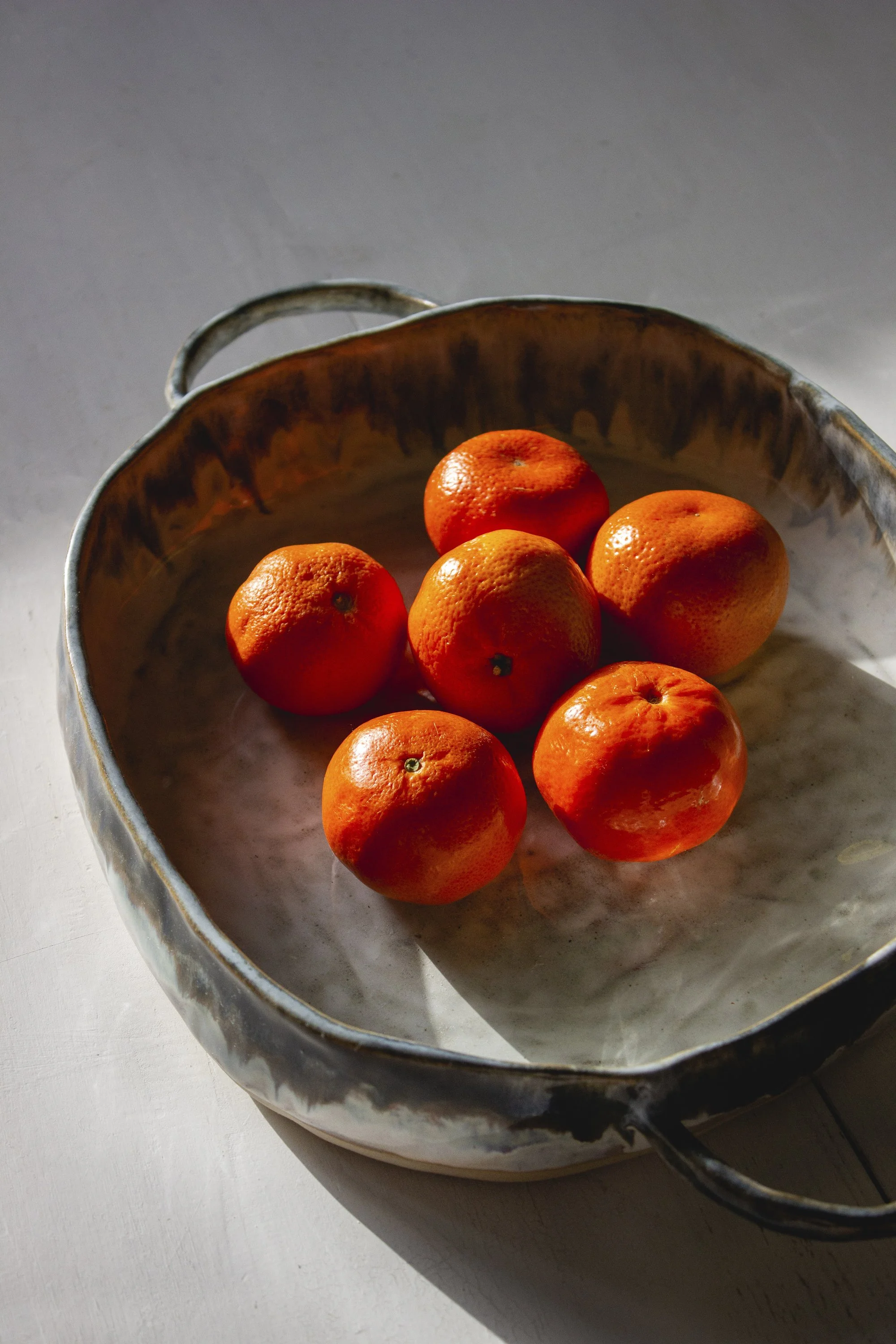 Kuvassa on seitsemän mandariinia laakeassa pyöreässä  astiassa.  Valo osuu kirkkaasti mandariineihin ja luo varjoja astian päälle.