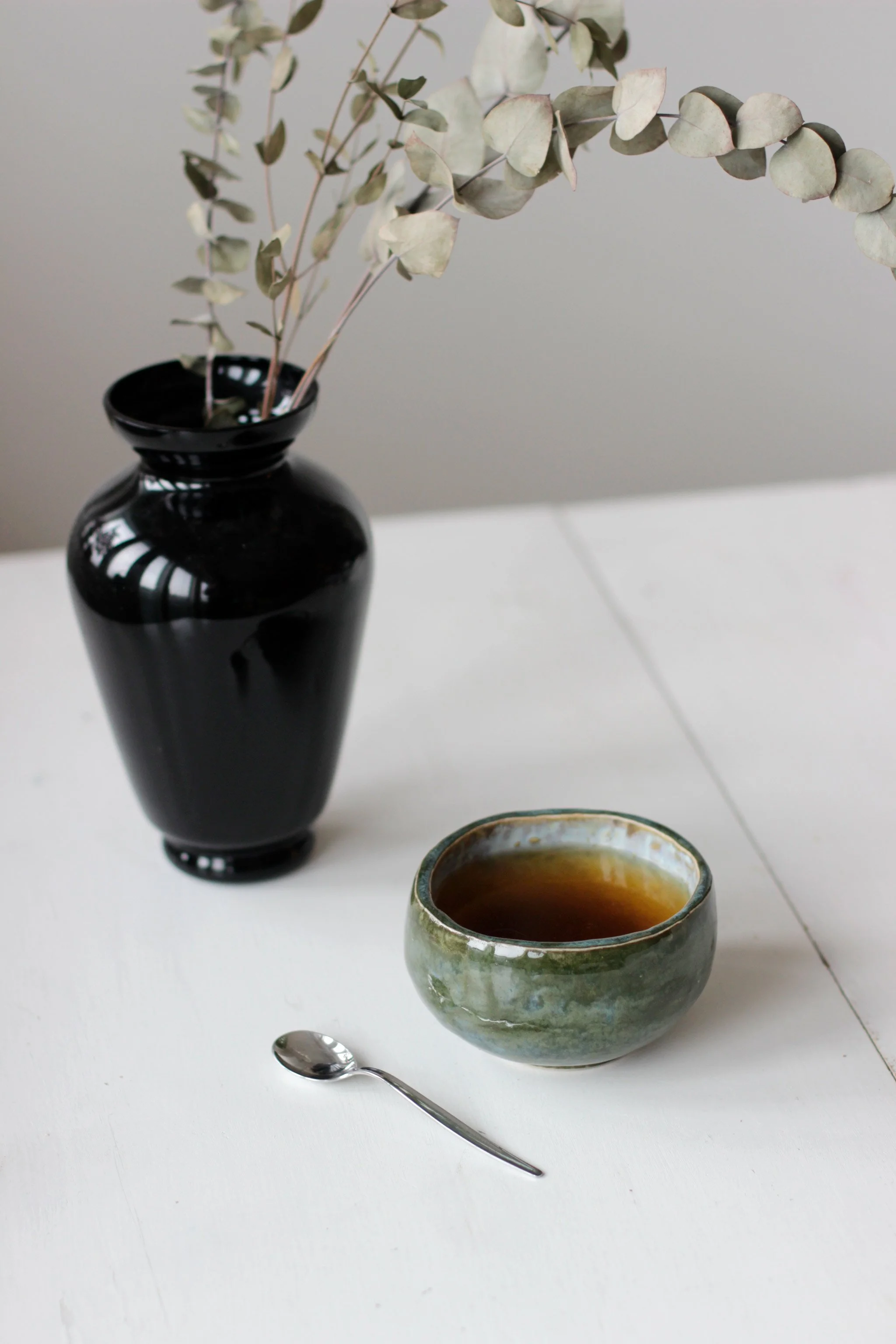 Mustavalkoinen pöytä, jossa keramiikkakulhossa tee, pieni hopeinen lusikka ja musta lasinen kukkaruukku, jossa kuivuneet eucalyptuslehdet.