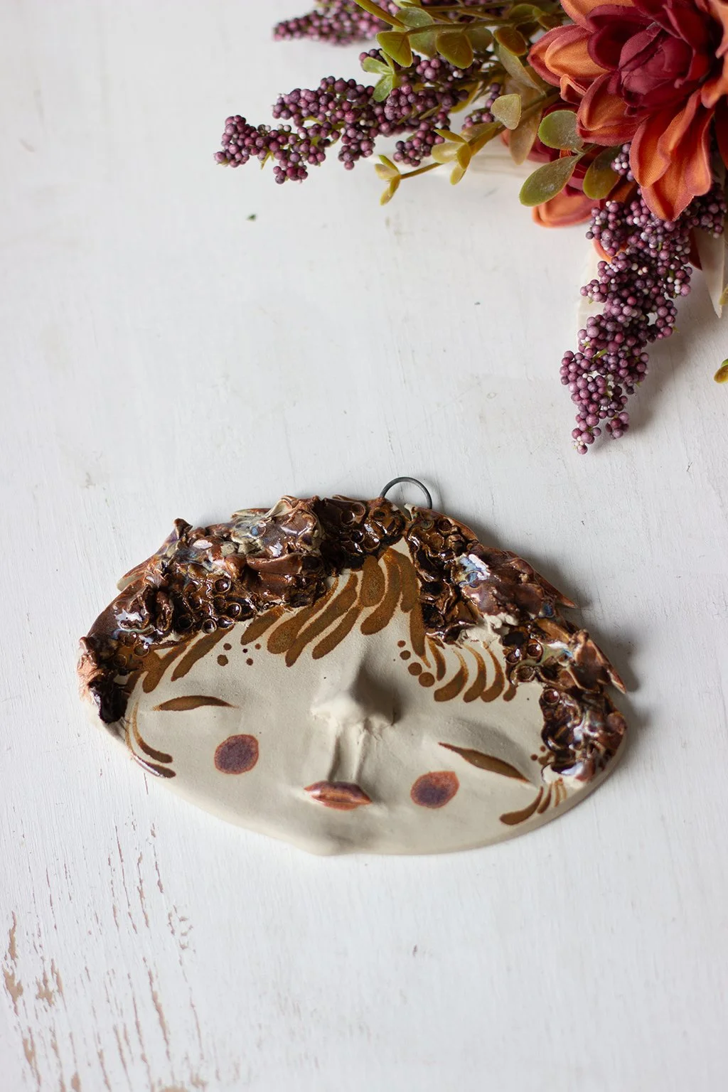 Kuva hienostuneesta kasvoreliefistä, jossa on koristeellisia yksityiskohtia. Taustalla kukkakimppu, lilja ja oranssinkeltaisia kukkia.